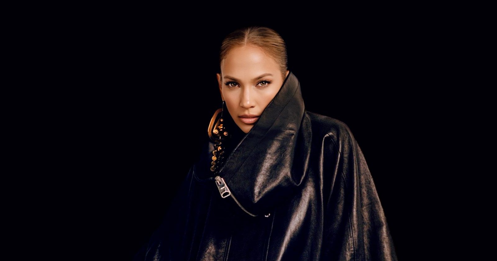 Jennifer Lopez vor schwarzem Hintergrund: sie trägt eine dunkelbraune, glänzende Jacke mit einem großen Zippverschluss und murmelförmige, lange Ohrringe