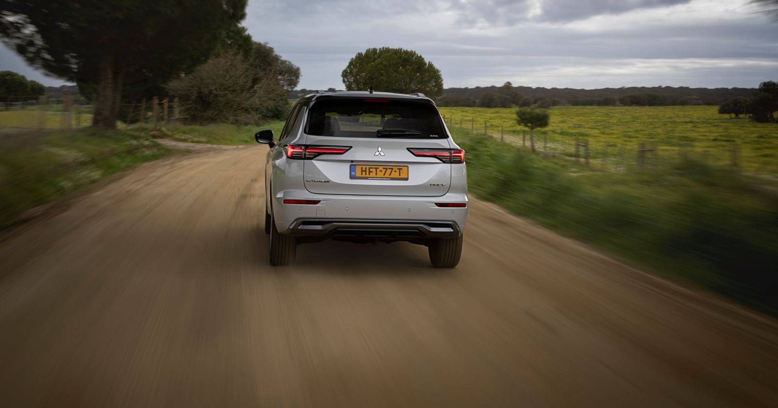 Das silberne Heck des Mitsubishi Outlander. Das Auto fährt auf eine braunen Schotterstraße. Links und rechts sind grüne Wiesen und einige Bäume zu sehen.