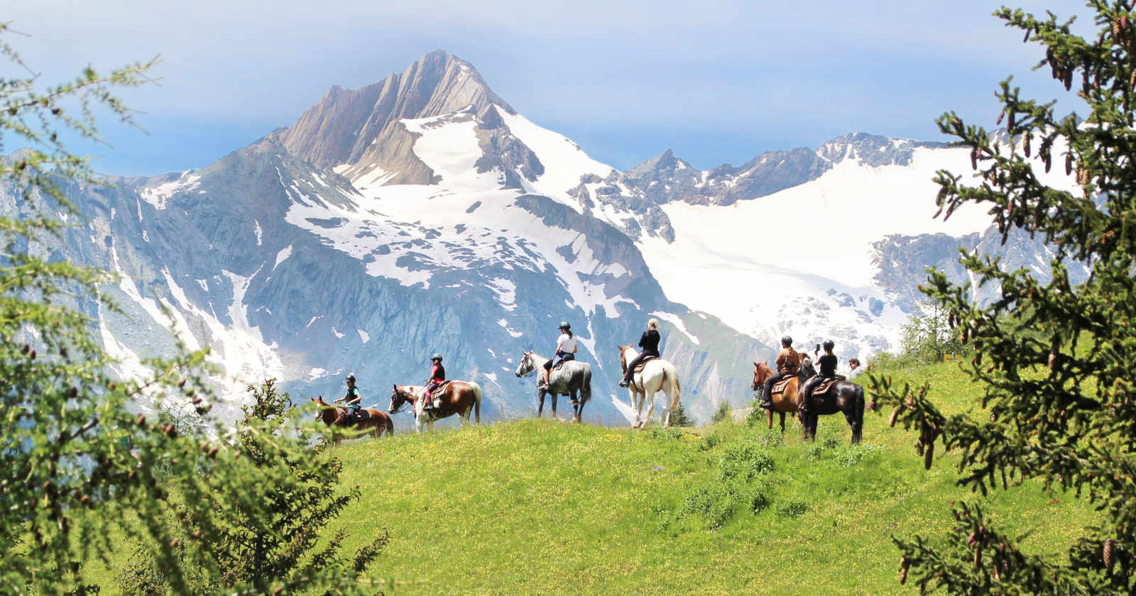 Menschen reiten über eine Wiese, dahinter sind Berge zu sehen