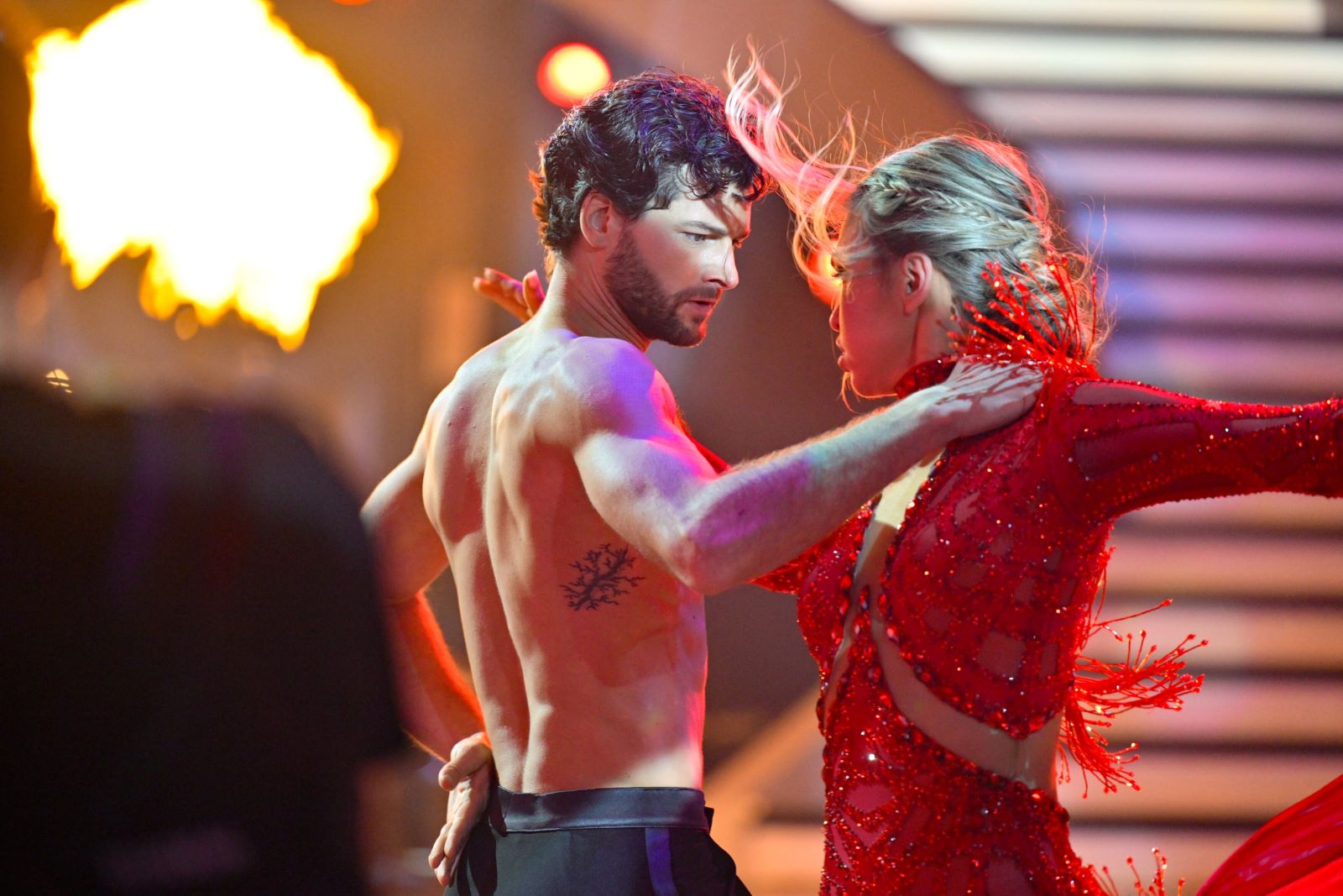 Aaron Karl tanzt im Finale von Dancing Stars 2025 einen Paso Doble ohne Hemd. Er steht eng mit Tanzpartnerin Katya Mizera, die ein rotes Glitzerkleid trägt. Im Hintergrund lodert eine Flamme.