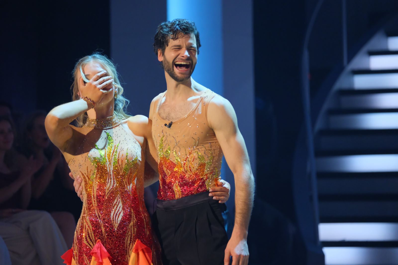Aaron Karl und Katya Mizera im Moment ihres Sieges bei Dancing Stars 2025. Beide tragen feuerfarbene Kostüme, Katya hält sich überrascht die Hand vors Gesicht, Aaron lacht.