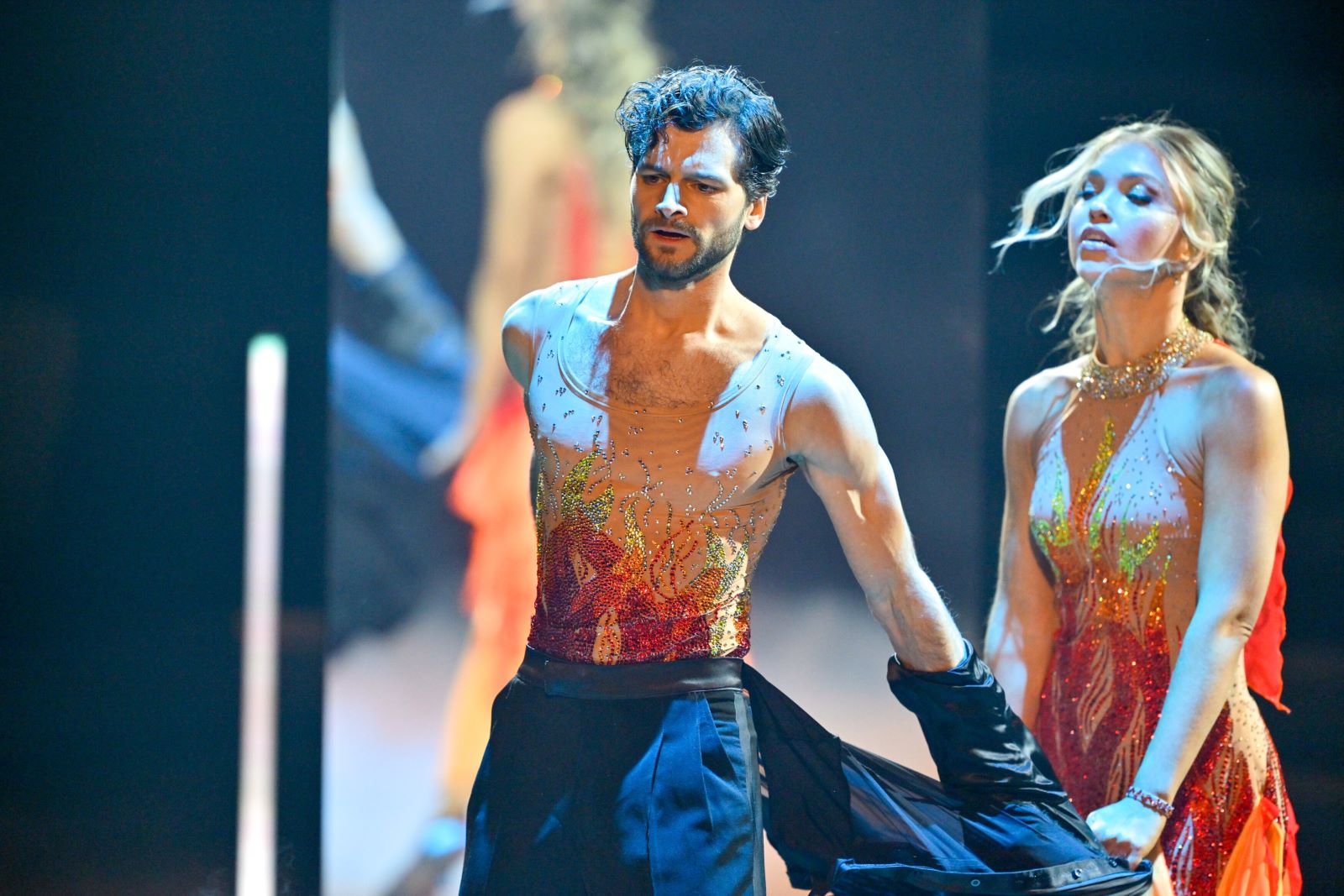 Aaron Karl steht im Dancing Stars-Finale 2025 mit geöffnetem Jackett auf der Bühne. Unter dem schwarzen Anzug trägt er ein hautenges, glitzerndes Top mit rot-goldenen Flammen. Katya Mizera steht neben ihm.
