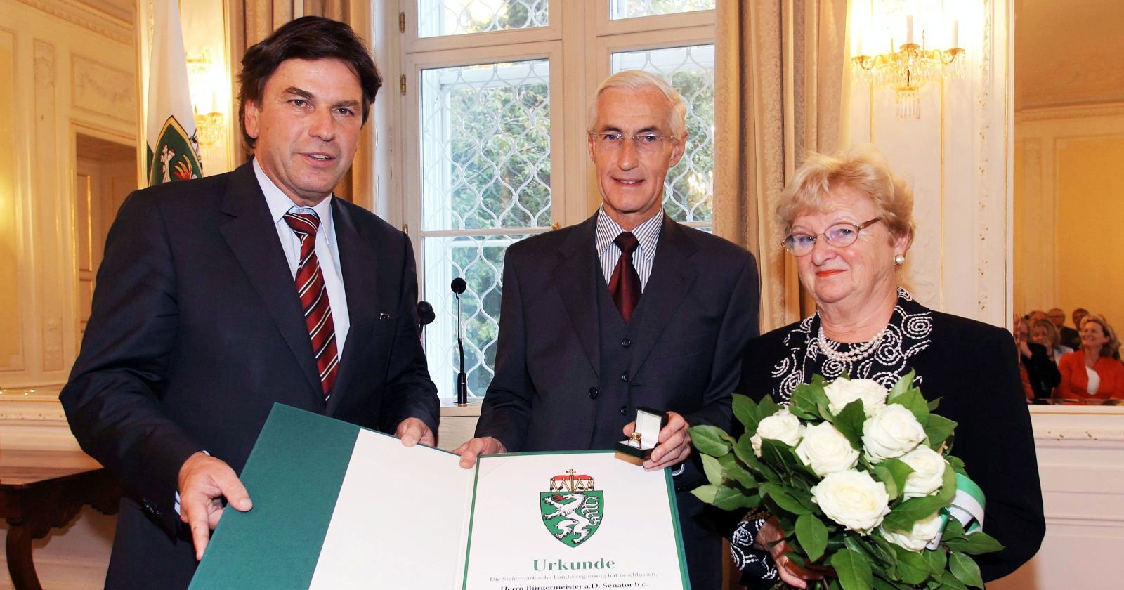 Zwei Männer und eine Frau stehen nebeneinander in einem festlichen Raum, der Mann in der Mitte bekommt eine Auszeichnung überreicht.