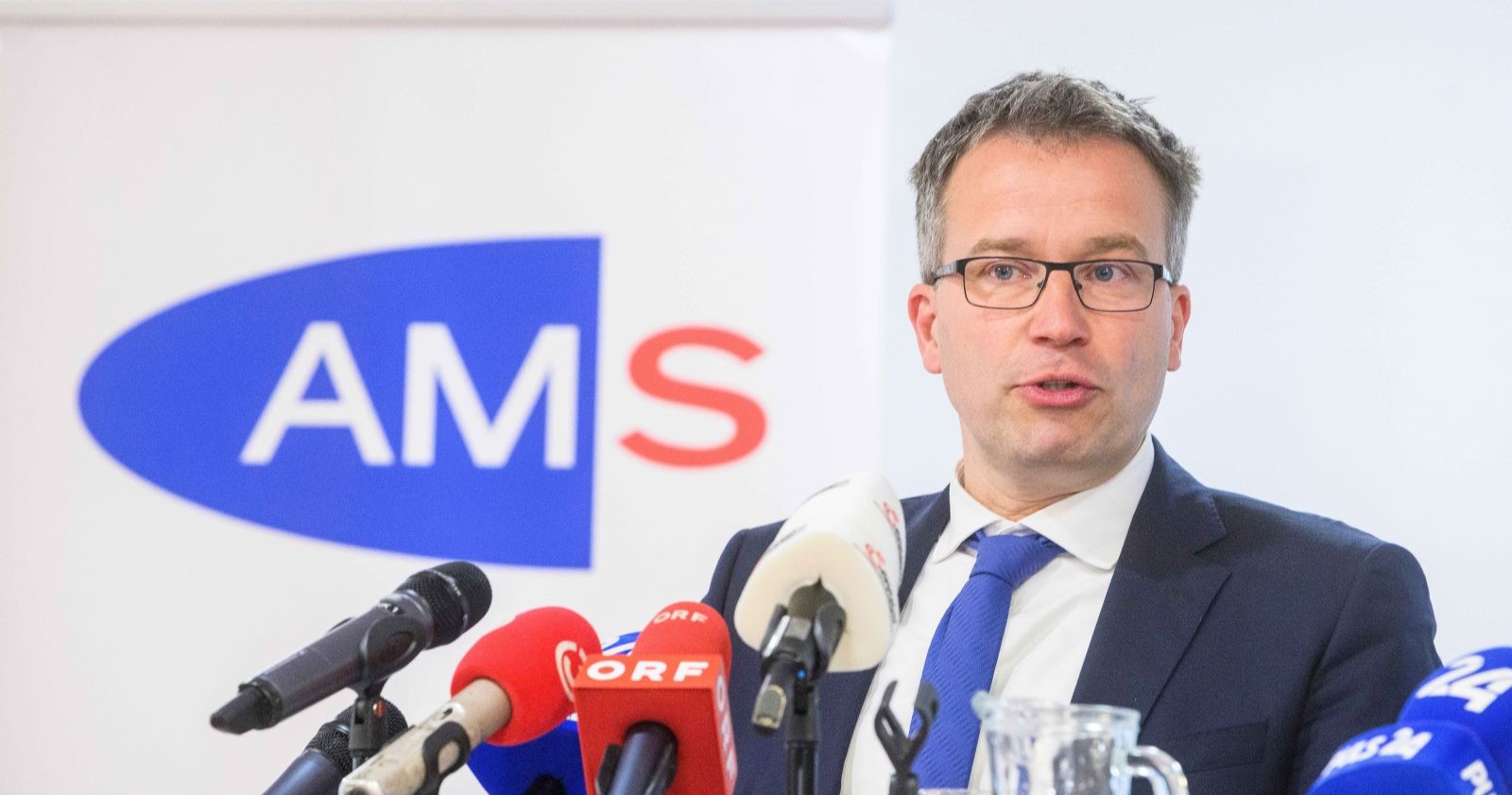 AMS-Chef Johannes Kopf bei einer Pressekonferenz vor Mikrofonen und dem Logo des Arbeitsmarktservice