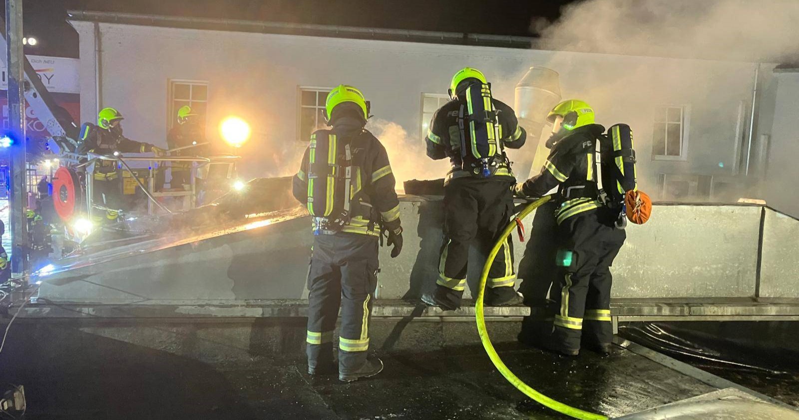 Vier Feuerwehrleute mit Atemschutzgeräten stehen nachts auf einem Flachdach im Einsatz gegen einen Brand; dichter Rauch steigt auf, ein Hubsteiger ist im Hintergrund im Einsatz.