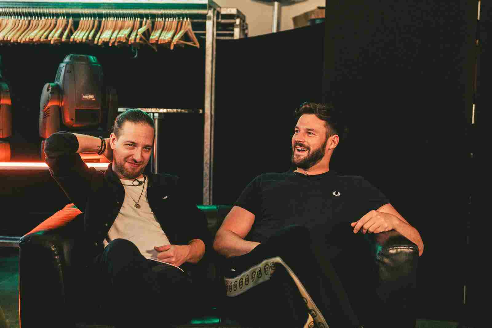 Daniel Fellner und Paul Pizzeria (Band &quot;Aut of Orda&quot;) sitzen entspannt und lächelnd gemeinsam auf einer Couch. Im Hintergrund ist eine Stange mit vielen Kleiderbügeln zu sehen, ansonsten alles schwarz.
