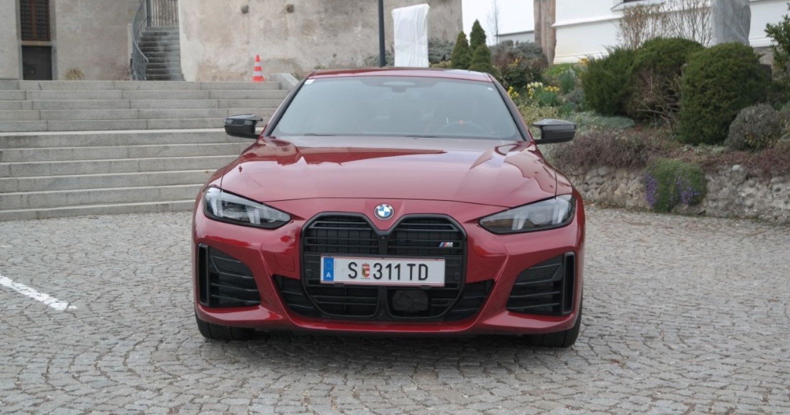 Ein weinroter BMW M440i steht auf einem Platz, der aus Pflastersteinen besteht. Im Hintergrund ist eine Steinstufe zu sehen, recht im Bild einige Büsche.