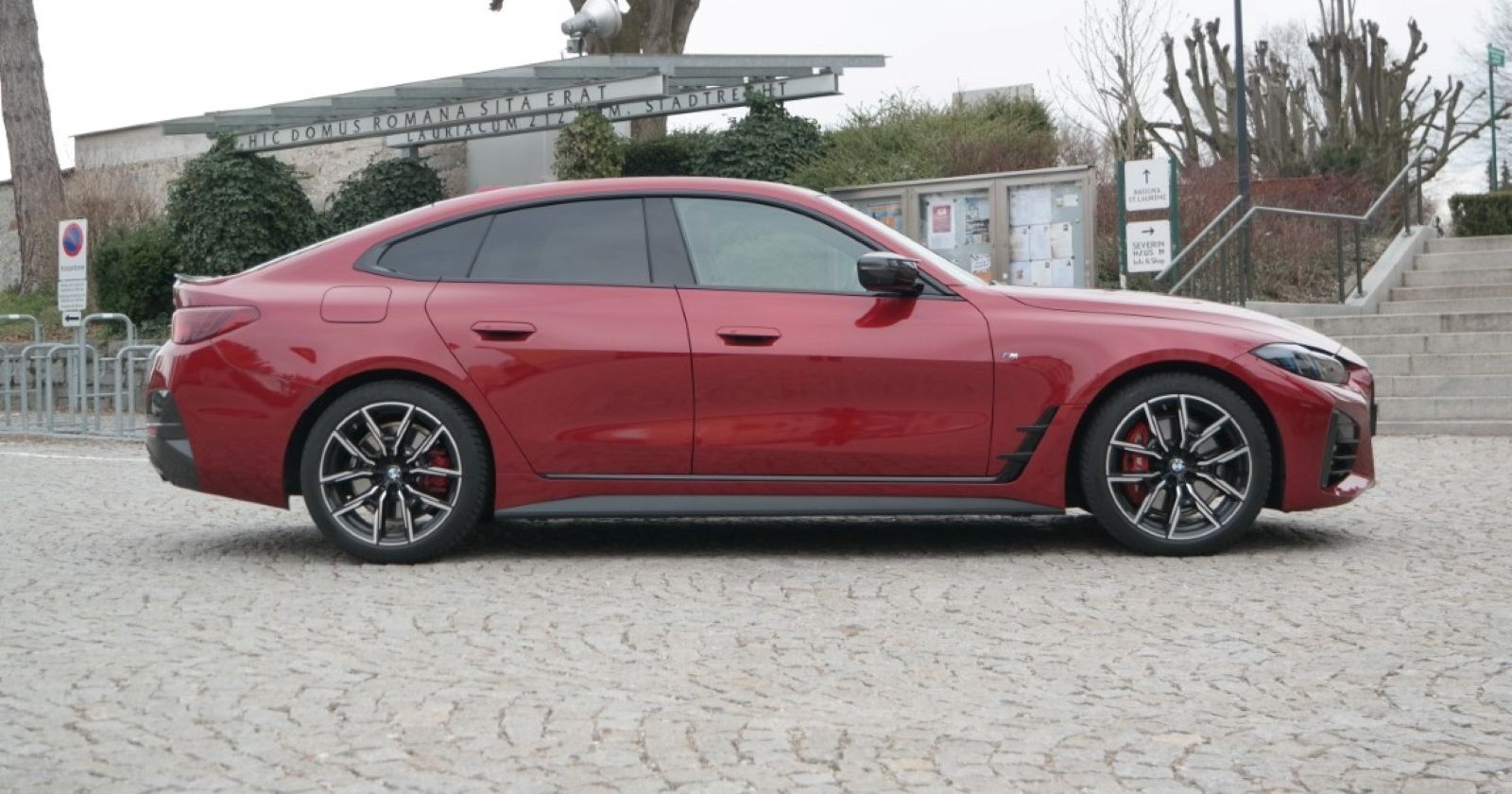 Ein weinroter BMW M440i von der Seite. Das Auto parkt auf einem Platz, der aus Pflastersteinen besteht. Im Hintergrund sind Bäume ohne Blätter und eine Stiege zu sehen.