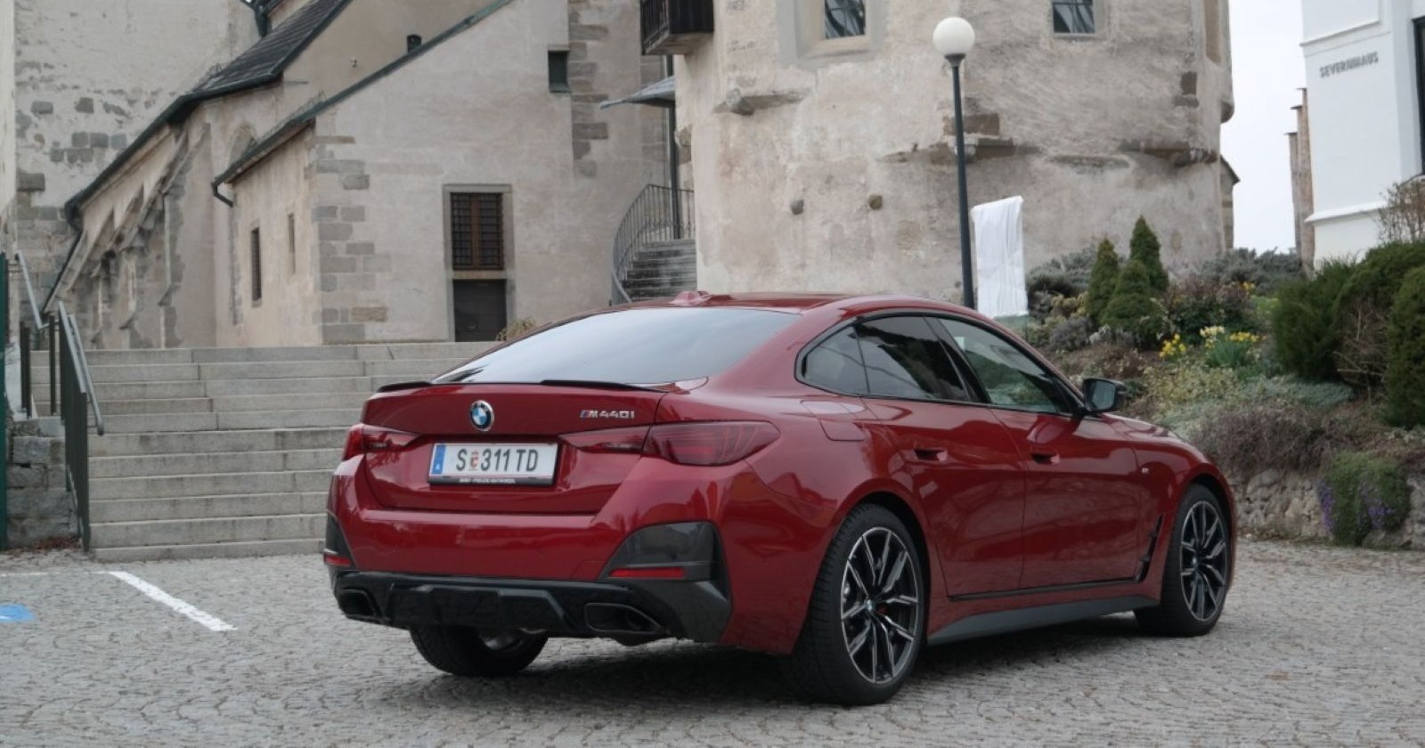 Ein weinroter BMW M440i parkt vor einer alten Kirche, die aus Steinen gebaut ist. Vor der Kirche sind einige Büsche und eine Steinstiege zu sehen.