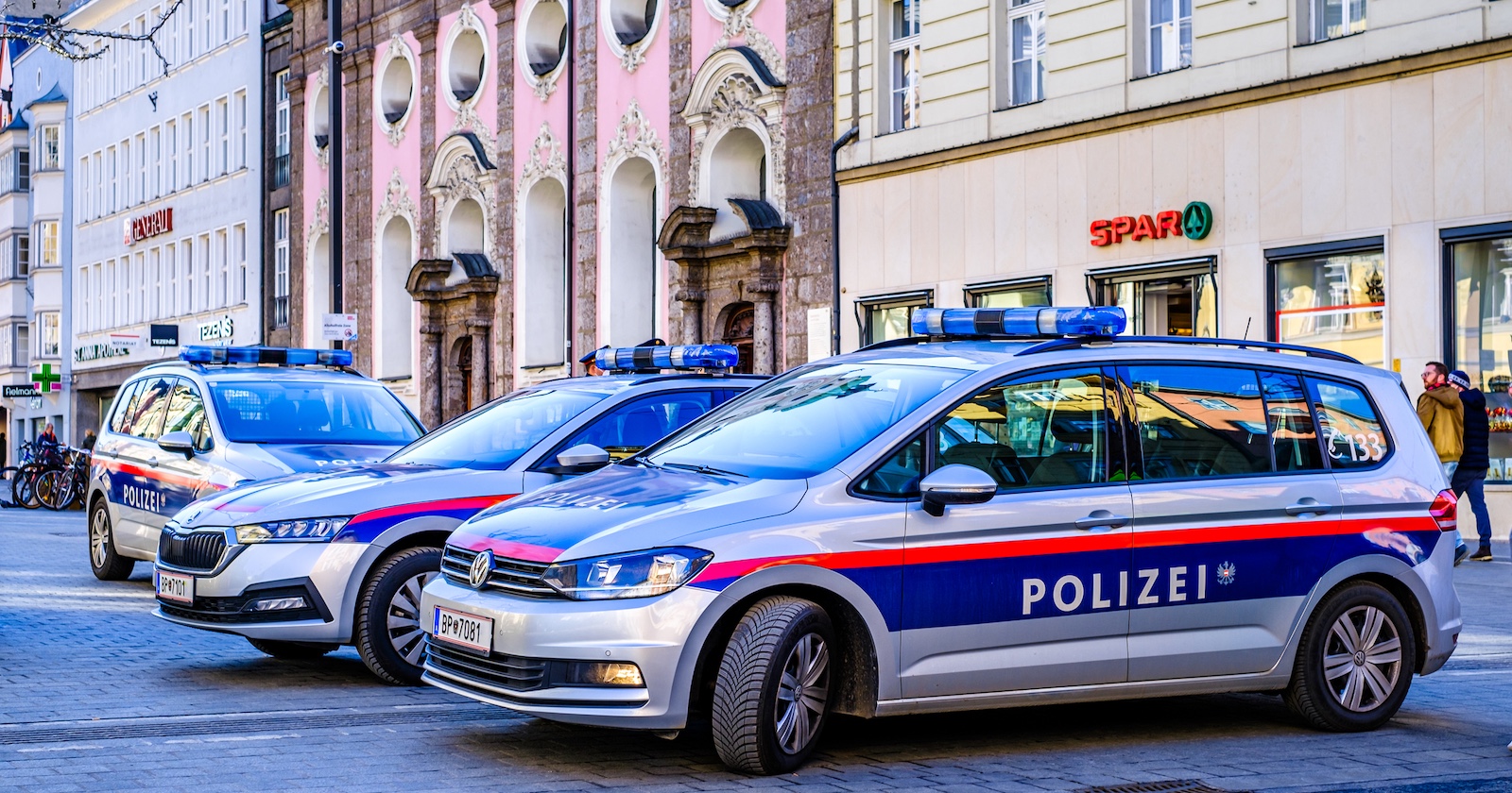Drei österreichische Polizeiautos mit Blaulicht stehen nebeneinander in einer Fußgängerzone in der Innsbrucker Innenstadt. Im Hintergrund sind ein SPAR-Supermarkt, Altbauten und Passanten zu sehen.