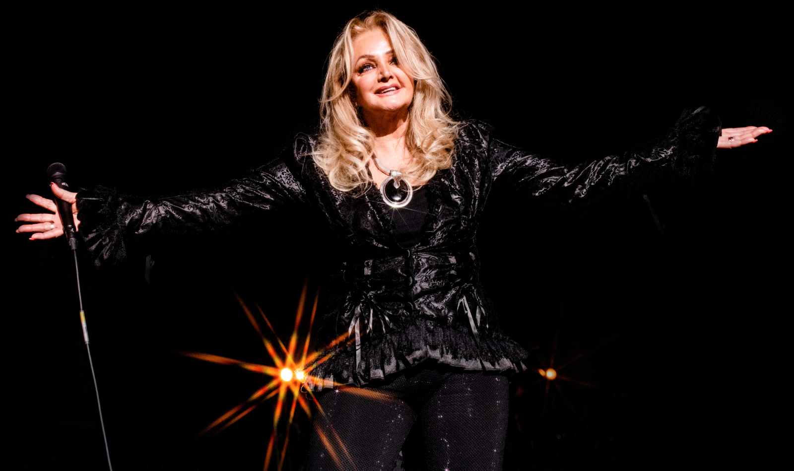 Bonnie Tyler auf der Bühne mit komplett schwarzem Outfit, die Hände seitlich ausgestreckt, in der einen Hand hält sie das Mikrofon.