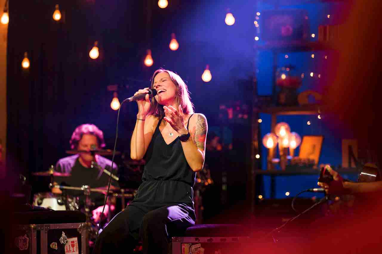 Sängerin Christina Christl Stürmer sitzt auf der Bühne und singt ins Mikrofon, im Hintergrund ist ein Schlagzeuger zu sehen und Glühbirnen.