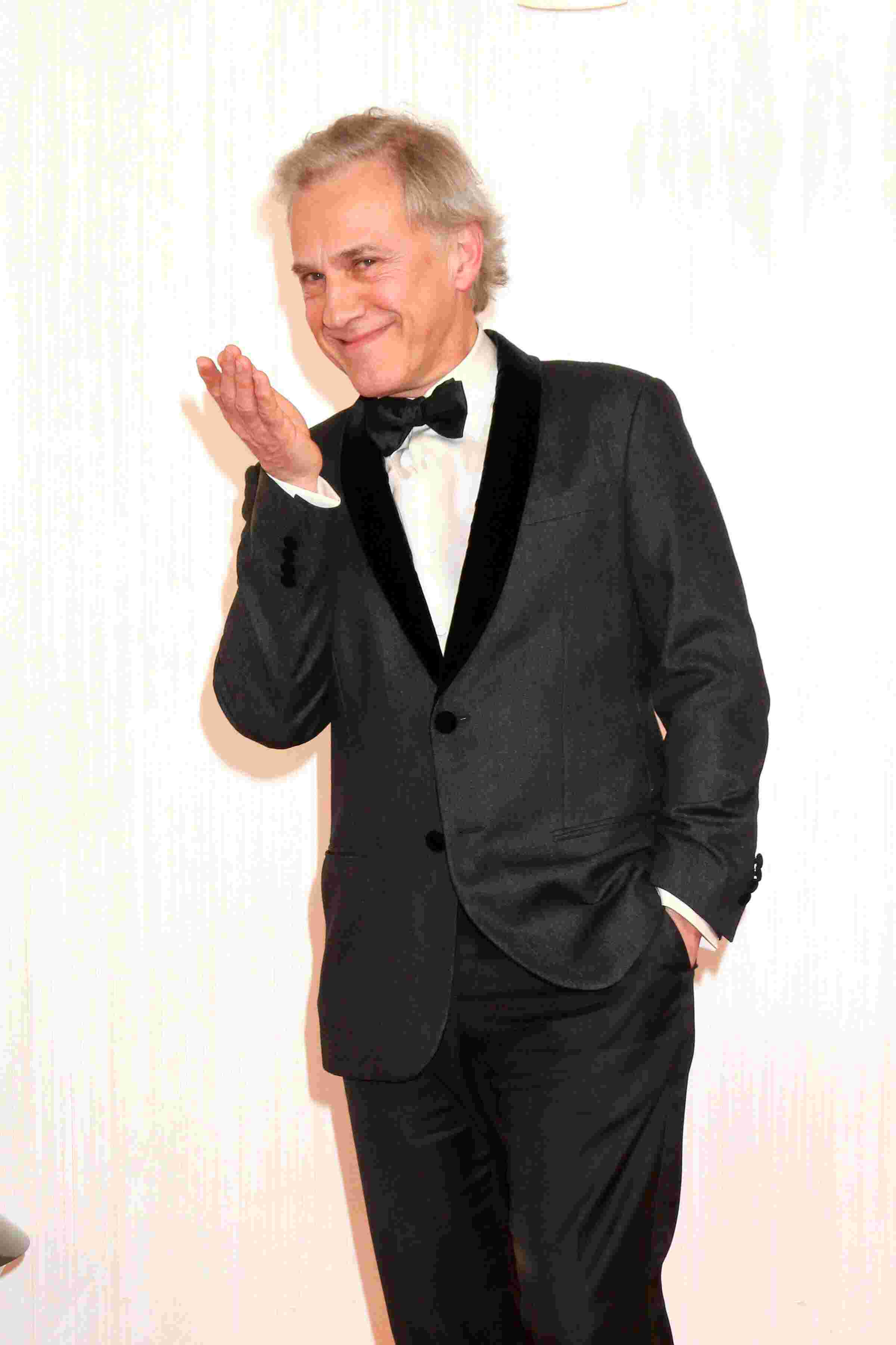 Christoph Waltz in schwarzem Smoking mit Fliege, lächelnd und eine Hand zum Kussmund geformt, vor hellem Hintergrund.