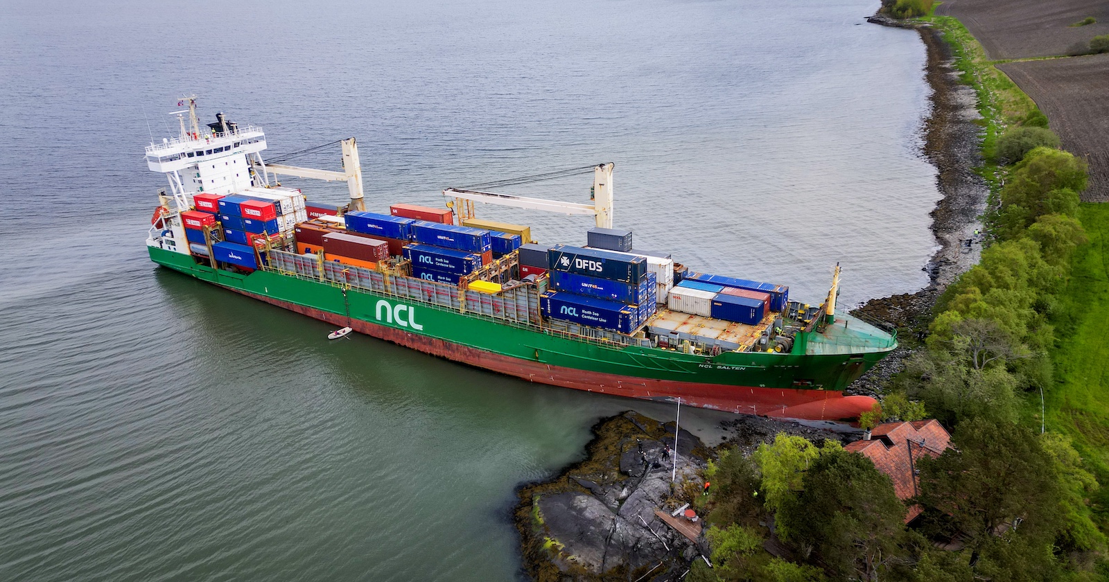Luftaufnahme eines grünen Containerschiffs mit der Aufschrift „NCL“, das im flachen Wasser eines Fjords in Norwegen gestrandet ist. Der Bug des Schiffs liegt direkt vor einem roten Holzhaus am Ufer, nur wenige Meter von der Gebäudewand entfernt.