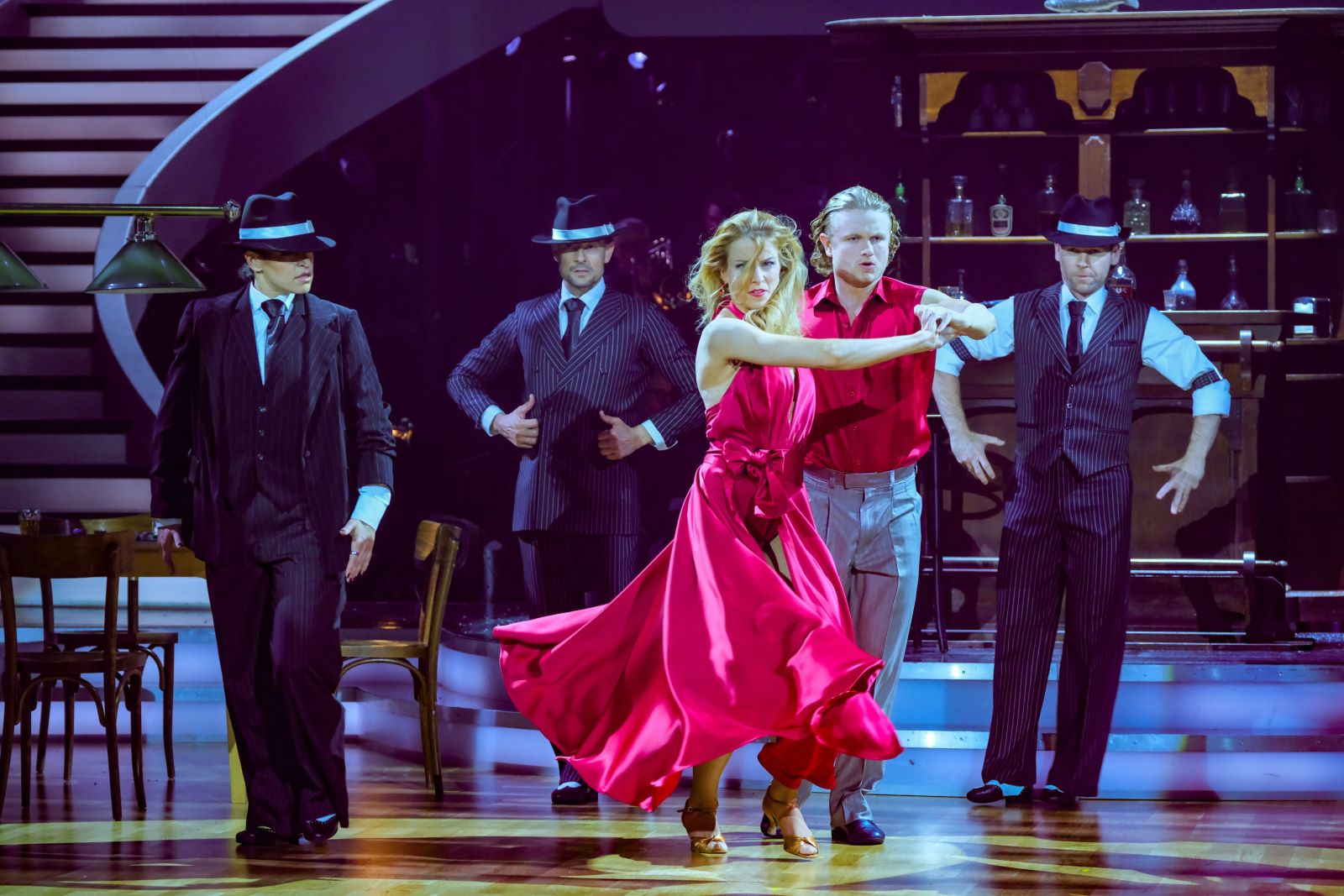 Paulus Bohl und Catharina Malek tanzen im Dancing Stars-Finale 2025 einen Showtanz im Mafia-Stil. Sie sind von Männern in Anzügen und Hüten umgeben, Catharina trägt ein rotes Kleid.