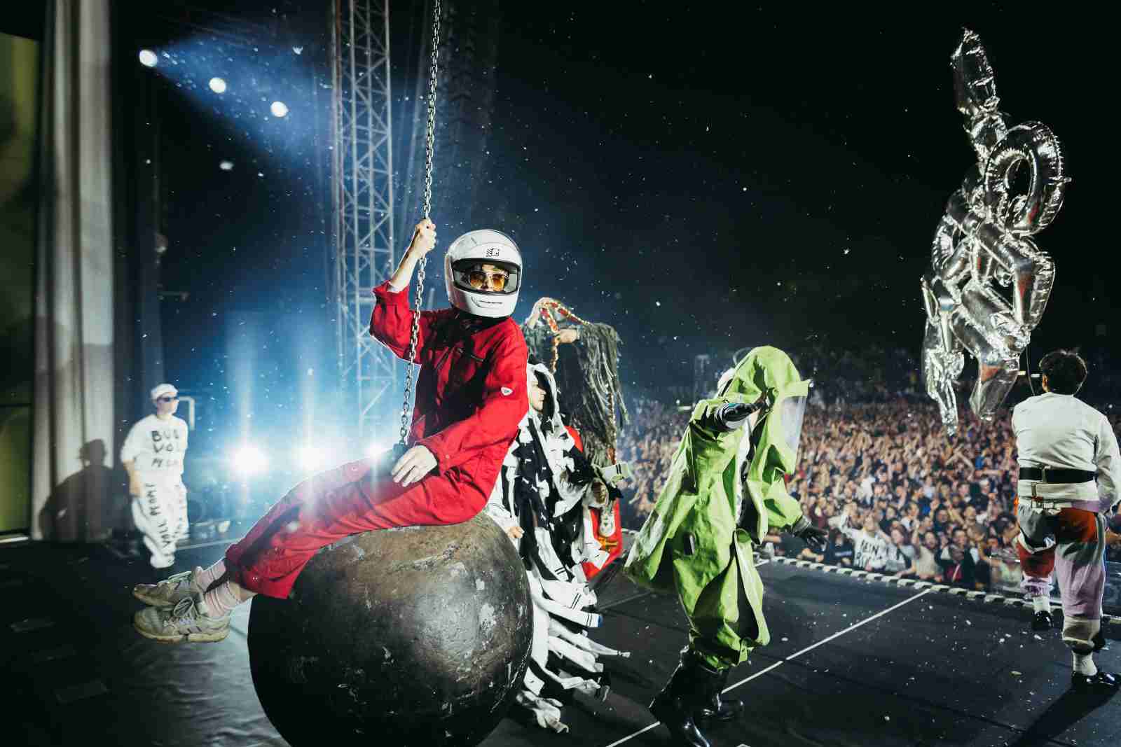 Die Band Deichkind bei einer spektakulären Bühnenshow. Im Vordergrund sitzt ein Mitglied in rotem Overall und Motorradhelm auf einer großen Abrisskugel, die an einer Kette hängt. Um ihn herum weitere Personen in schrillen Kostümen, darunter ein grüner Ganzkörperanzug. Im Hintergrund sieht man eine große, jubelnde Menschenmenge und helle Scheinwerferlichter, die die Szene dramatisch ausleuchten. Rechts schwebt ein silberner Luftballon in Form einer Hand mit ausgestrecktem Zeigefinger.