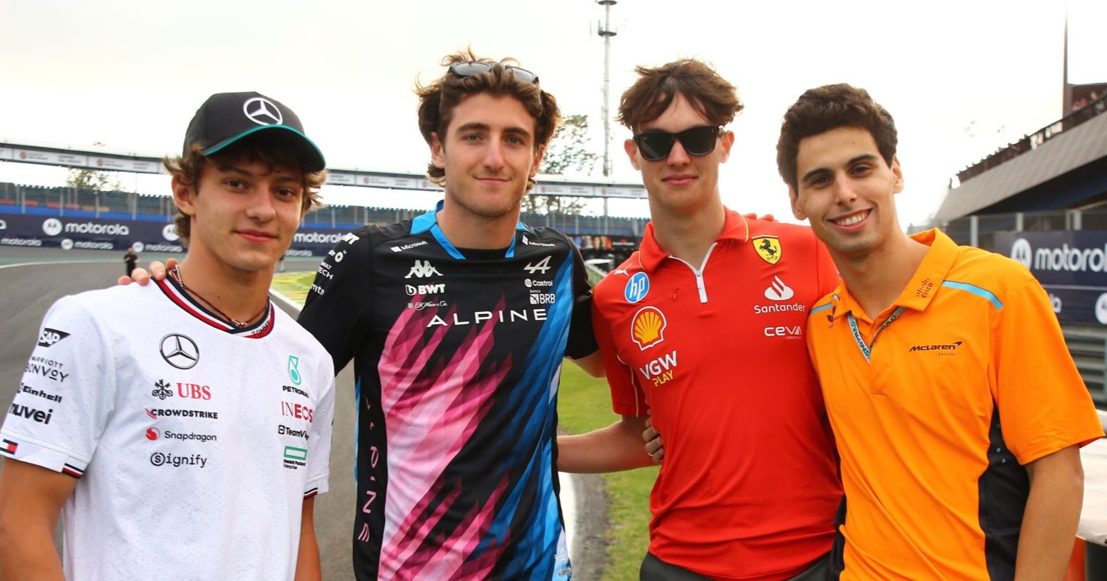 Vier junge Formel-1-Fahrer in Teamkleidung von Mercedes, Alpine, Ferrari und McLaren posieren gemeinsam an der Strecke.