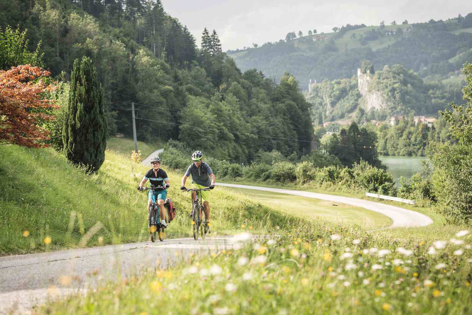 Paar fährt mit dem Fahrrad den Ennsradweg entlang, umgeben von Wäldern und Wiese, im Hintergrund ist die Burgruine Losenstein zu erkennen.