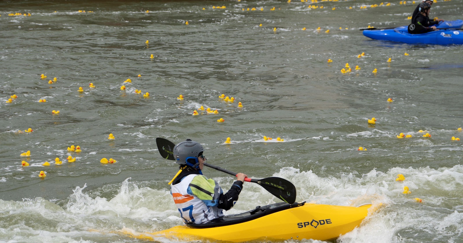 Wassersportler in einem gelben und einem blauen Kajak in einem Fluß in dem viele gelbe Plastikenten treiben