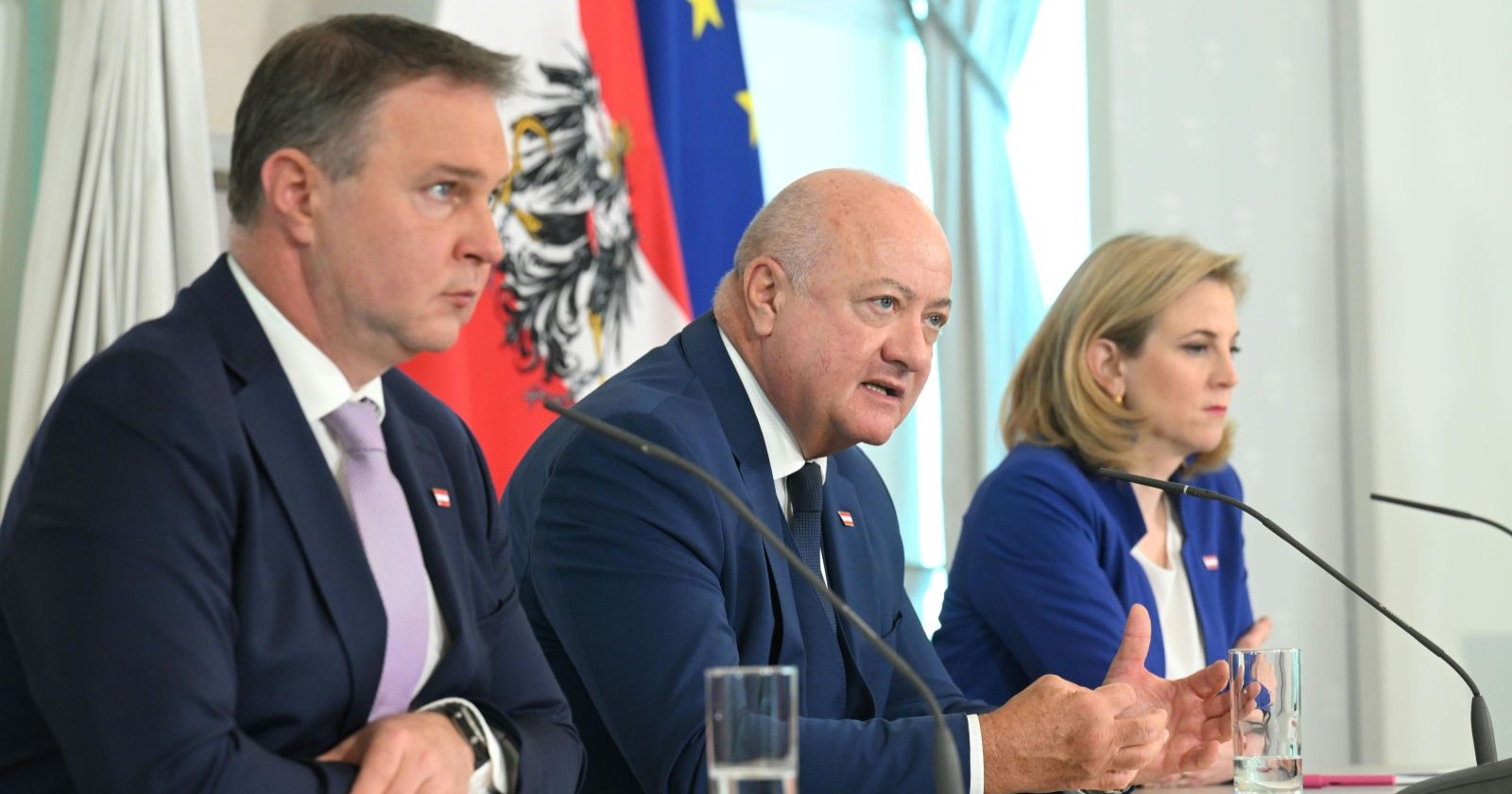 Andreas Babler, Christian Stocker und Beate Meinl-Reisinger sitzen bei einer Pressekonferenz vor Mikrofonen. Thema ist die Familienbeihilfe, konkret die Erhöhung und ausbleibende Valorisierung. Im Hintergrund sind österreichische und EU-Flagge sichtbar.