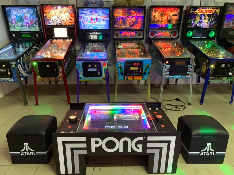 Ein Raum mit einer Sammlung klassischer Flipperautomaten an der Wand, darunter Titel wie &quot;Alice Cooper's Nightmare Castle&quot;, &quot;The Addams Family&quot;, &quot;Star Wars&quot; und &quot;The Machine: Bride of Pin·Bot&quot;. Im Vordergrund steht ein moderner, beleuchteter Atari PONG-Tisch mit zwei schwarzen Hockern, ebenfalls mit Atari-Logo. Der Raum ist gut beleuchtet und vermittelt den Eindruck eines Retro-Spielhallenambientes.