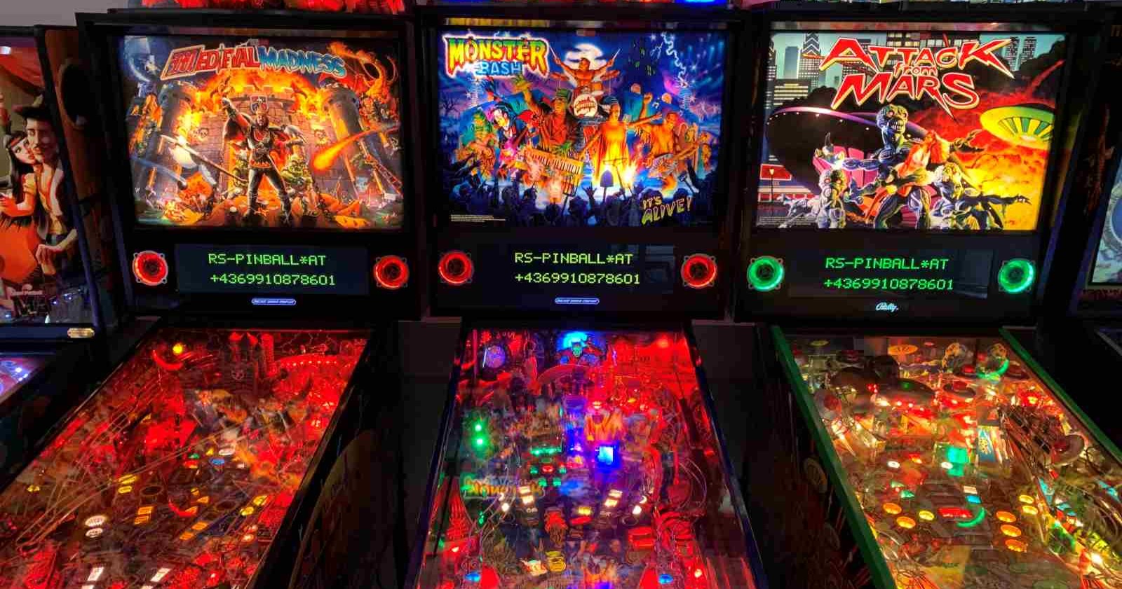 Drei beleuchtete Flipperautomaten nebeneinander in einer Spielhalle. Die Titel der Maschinen sind von links nach rechts: &quot;Medieval Madness&quot;, &quot;Monster Bash&quot; und &quot;Attack from Mars&quot;. Die Spielflächen sind bunt beleuchtet und zeigen jeweils thematisch passende Designs und Figuren.