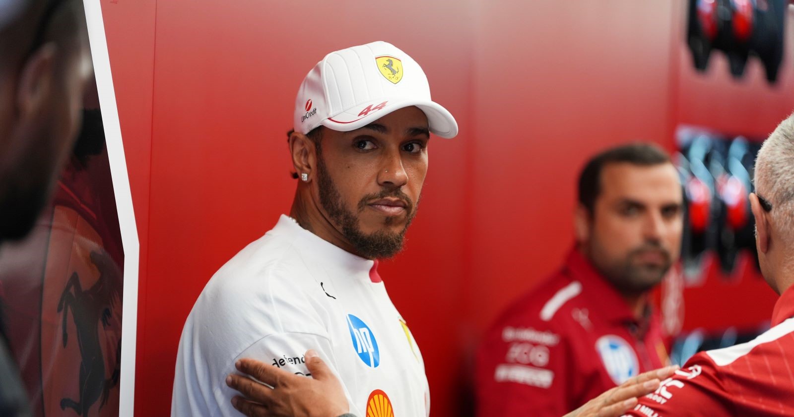 Lewis Hamilton in Ferrari-Teamkleidung steht mit ernster Miene in der Box, umgeben von Teammitgliedern. Deutlich sichtbar: das Ferrari-Logo im Hintergrund.