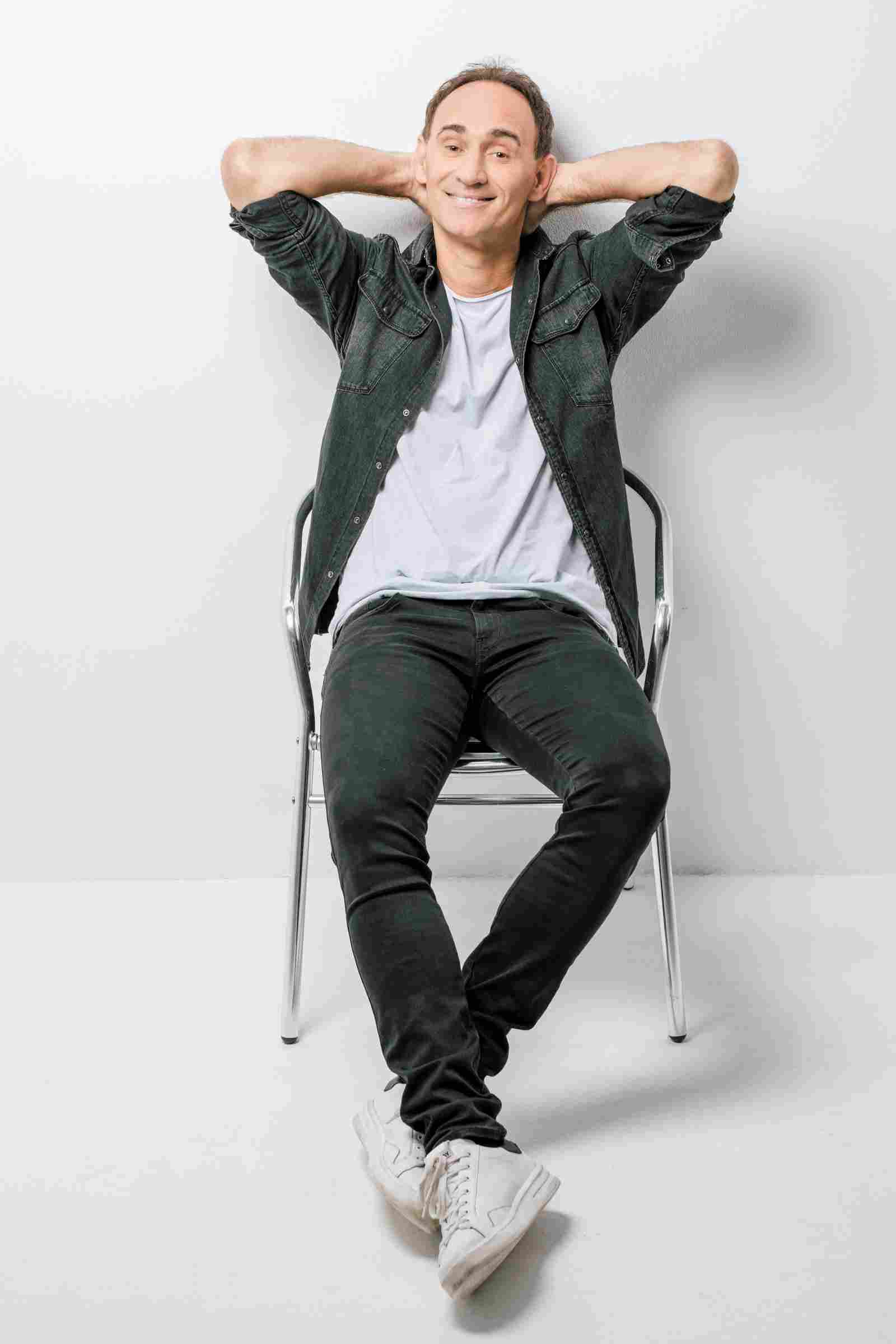 Ö3-Callboy Gernot Kulis sitzt lässig auf einem Stuhl, die Arme hinter dem Kopf verschränkt und lächelt.