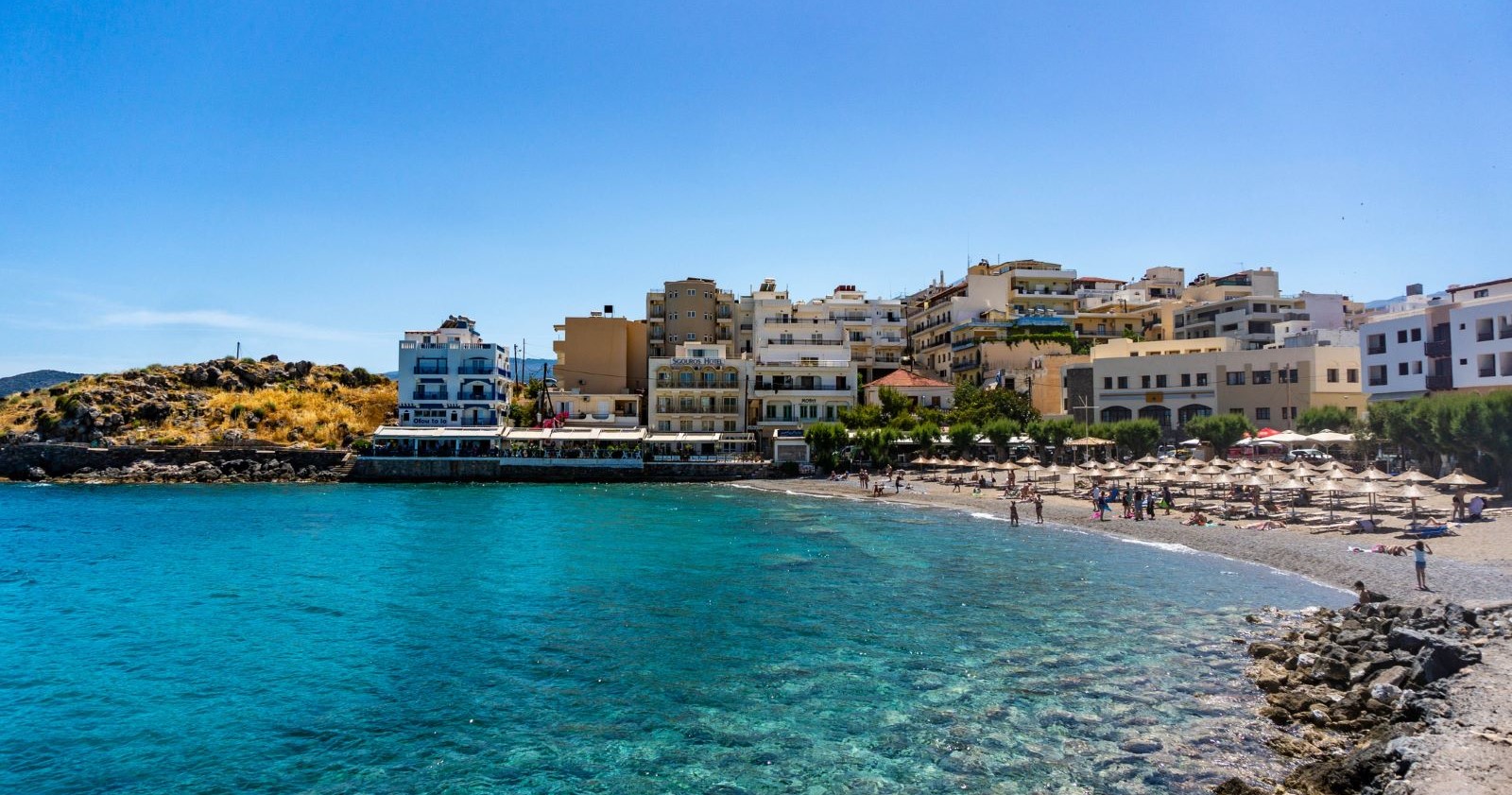 Strandpromenade von Agios Nikolaos auf Kreta mit türkisblauem Meer, Sonnenschirmen und Hotelgebäuden im Hintergrund bei klarem Himmel. Das Erdbeben in Griechenland hat keine größeren Schäden hinterlassen.