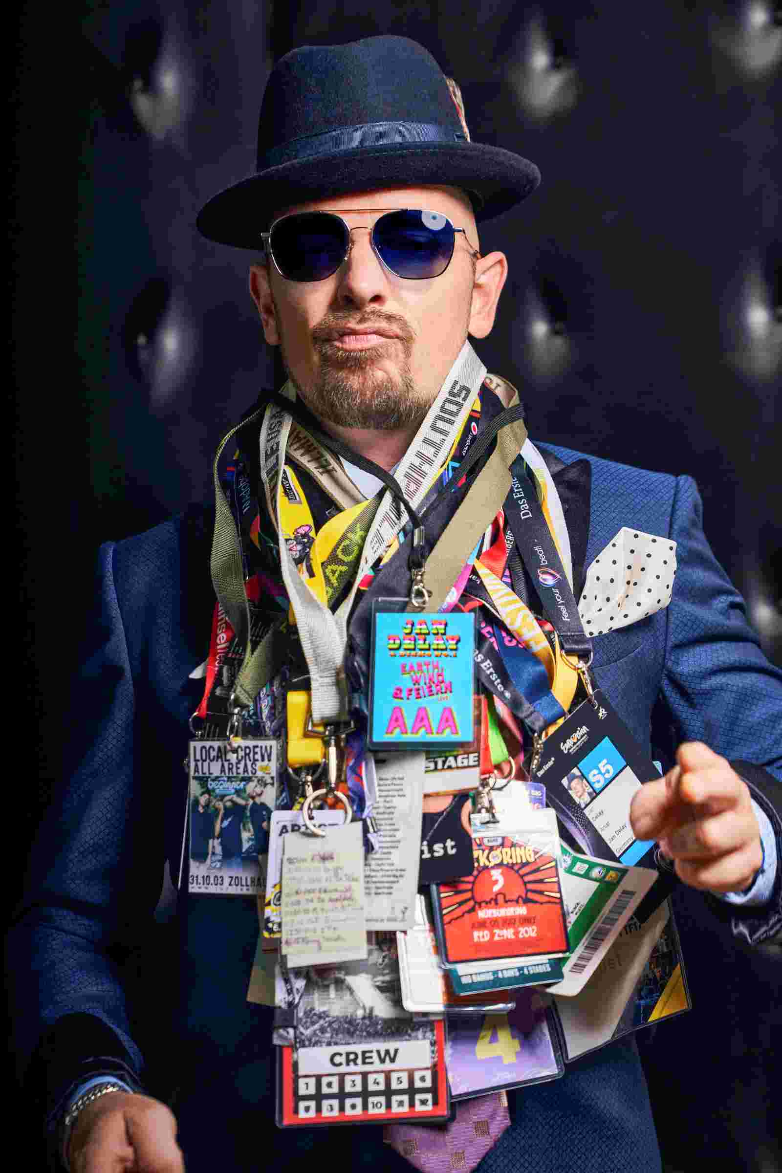 Musiker Jan Delay in dunkelblauem Sakko, Hut und Sonnenbrille. Um seinen Hals hängen mehrere Konzertpässe, sein Mund ist zu einem Duck Face geformt.