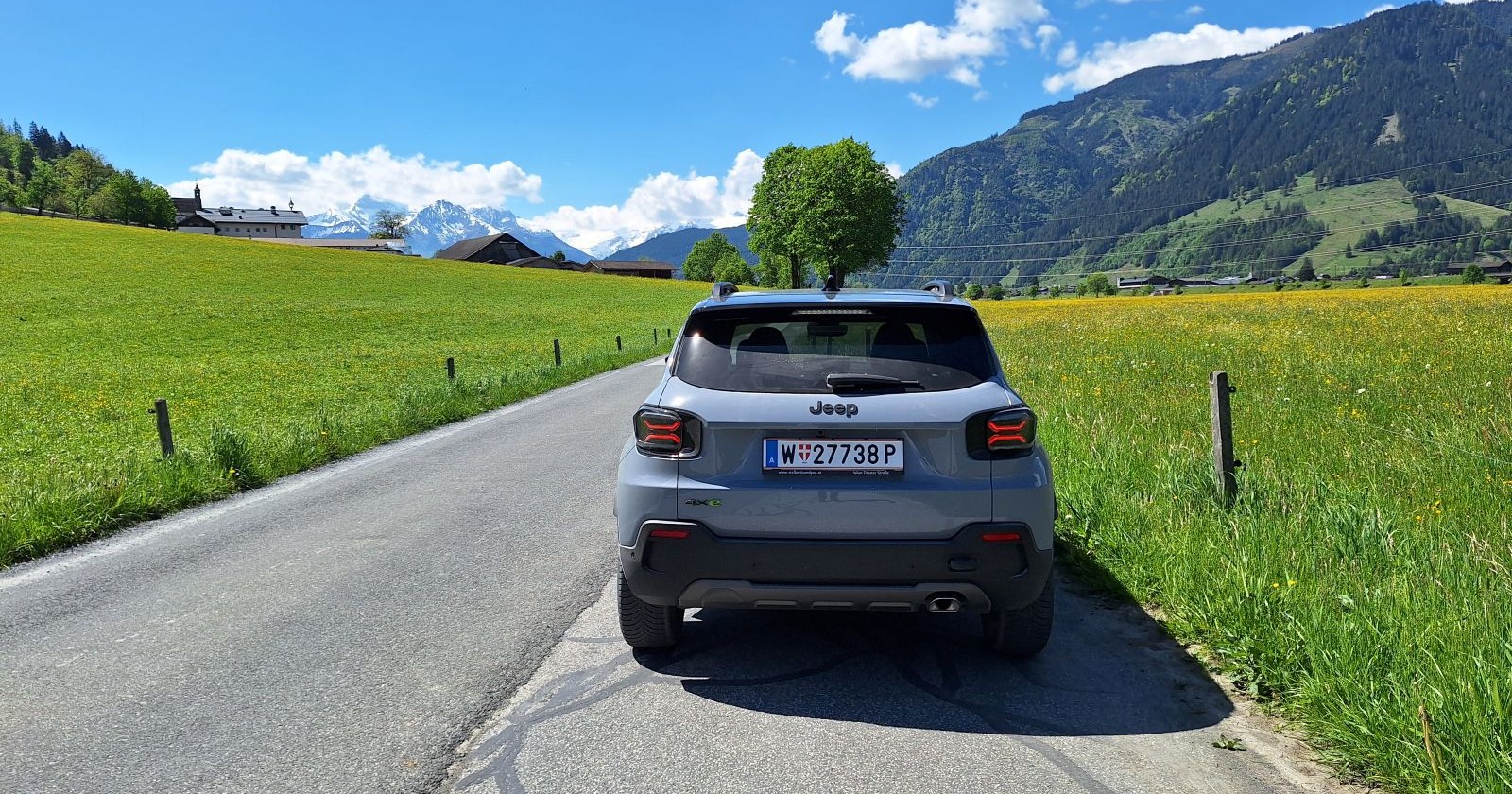 Das Heck eines blauen Jeep Avanger. Das Auto parkt auf eine einsamen Landstraße. Links und rechts ist eine Wiese zu sehen, im Hintergrund mittelhohe Berge, die bewaldet sind.