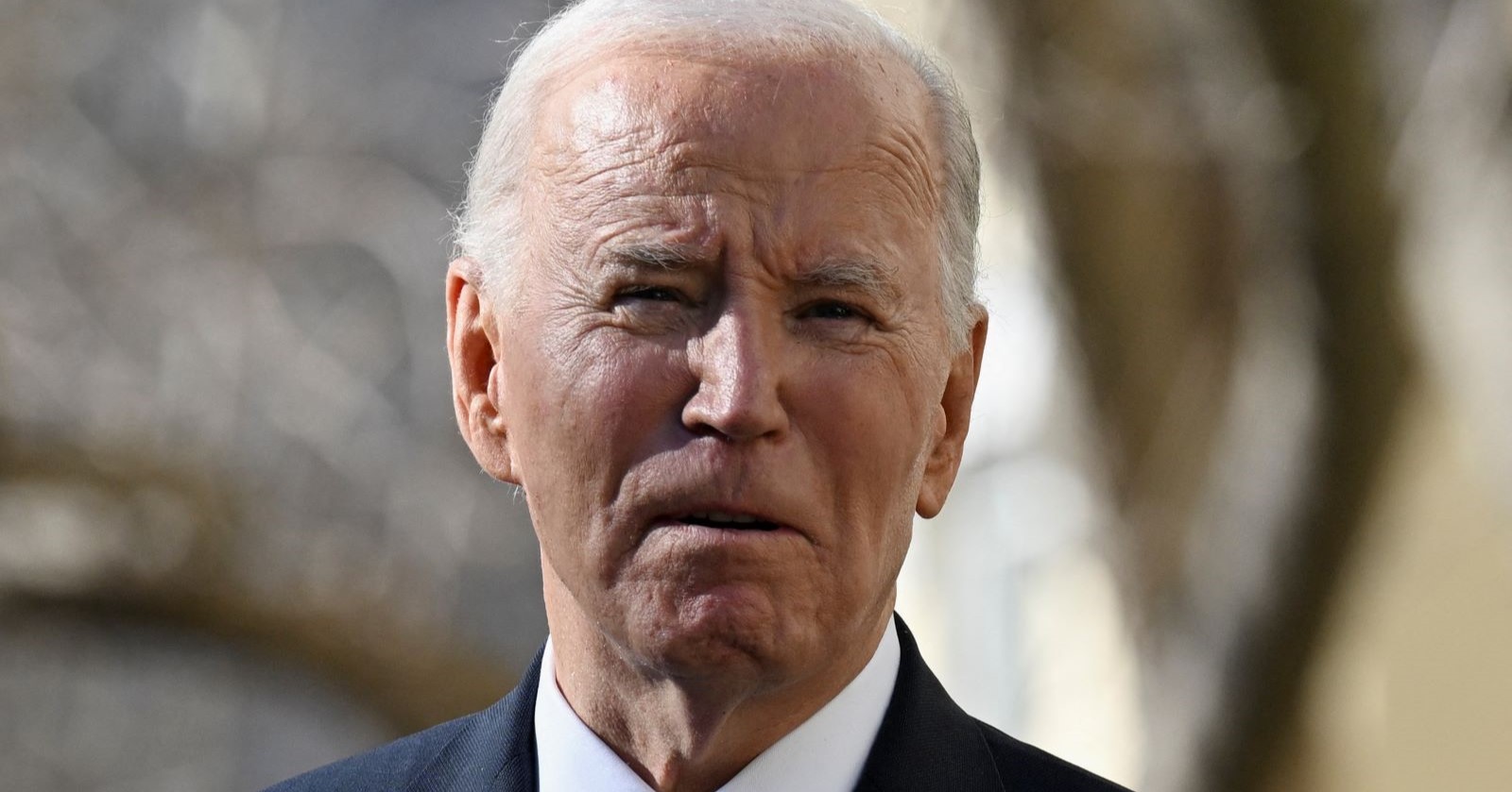 Porträt des krebskranken Joe Biden im Anzug, mit ernster Miene, vor unscharfem Hintergrund im Freien.