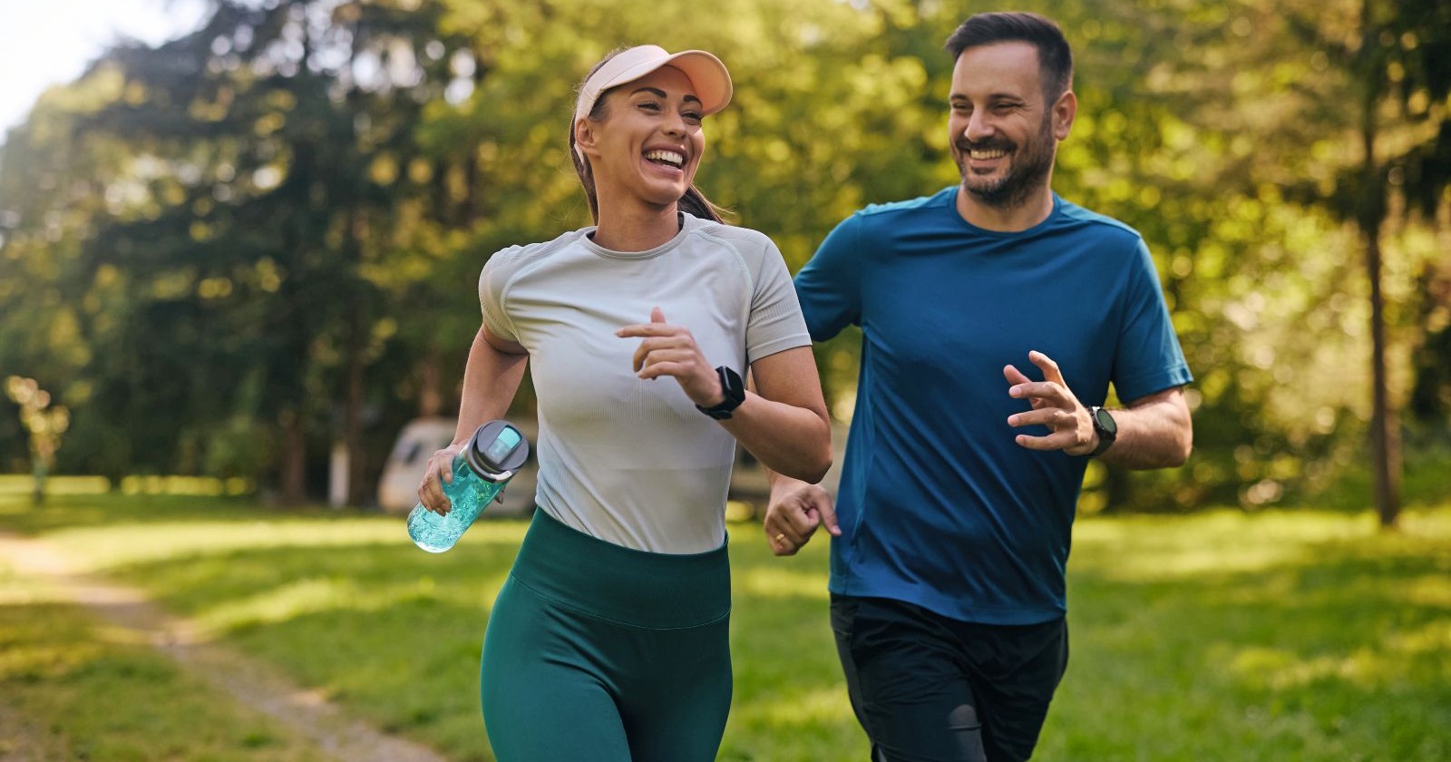 Eine Frau und ein Mann joggen. Beide lachen. Sie hat eine grüne Hose und ein weißes T-Shirt an, zudem trägt sie eine Schirmkappe. In einer Hand hält sie eine grüne Trinkflasche. Der Mann mit Vollbart hat eine blaues T-Shirt und eine schwarze Jogging-Hose an. Im Hintergrund sind Bäume und eine Wiese zu sehen.