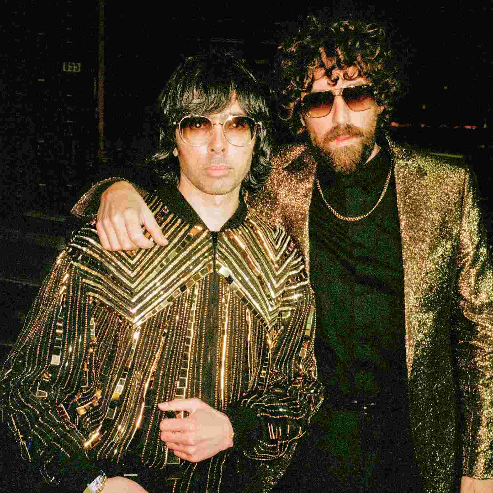 Die Band Justice - zwei Männer in auffalendem Outfit aus gold und schwarz, mit Sonnenbrillen, der eine hat den Arm über den anderen geschlagen. Beide schauen cool gerade in die Kamera.