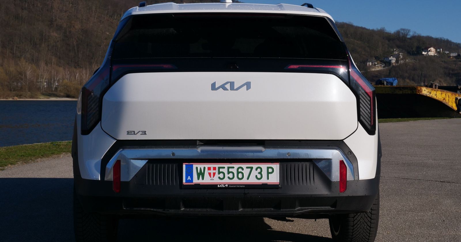 Das Heck eines weißen KIA EV3 leuchtet in der Sonne. Das Auto parkt am Rande eines Gewässers. Am Ufer ist ein Frachtschiff zu sehen.