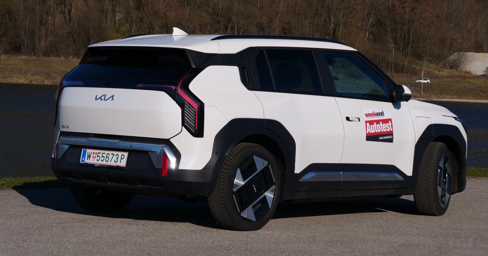 Ein weißer KIA EV3 von der Seite. Der Koreaner parkt am Ufer eines Gewässers. Am anderen Ufer fährt ein weißes Auto, zudem ist ein Wald zu sehen.