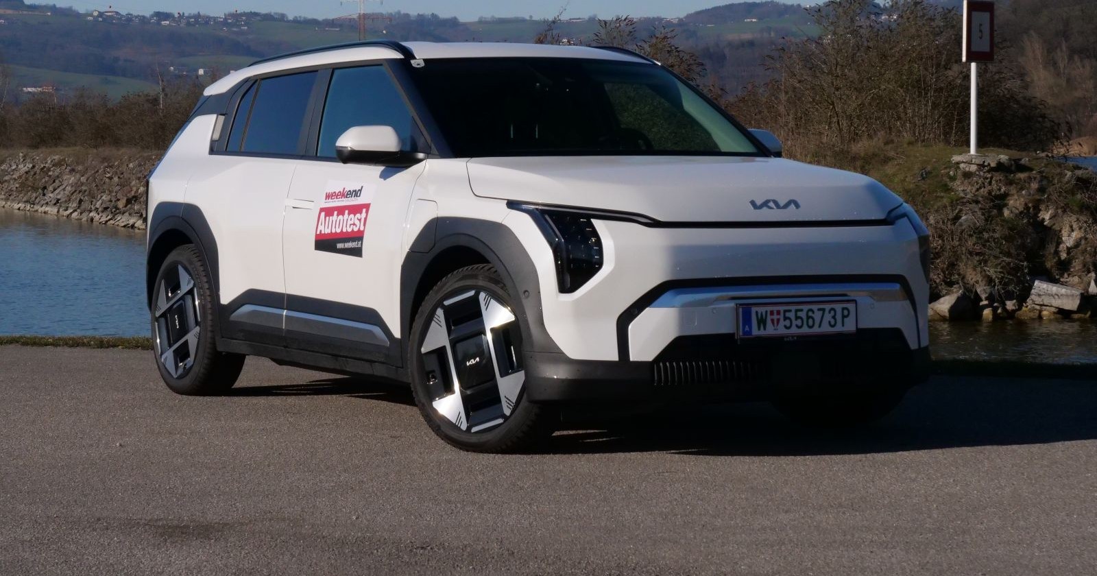 Ein weißer KIA  EV3 parkt auf einem asphaltieren Parkplatz. Das Auto wird von der Sonne angeleuchtet, im Hintergrund sind ein Gewässer und Hügel zu sehen.