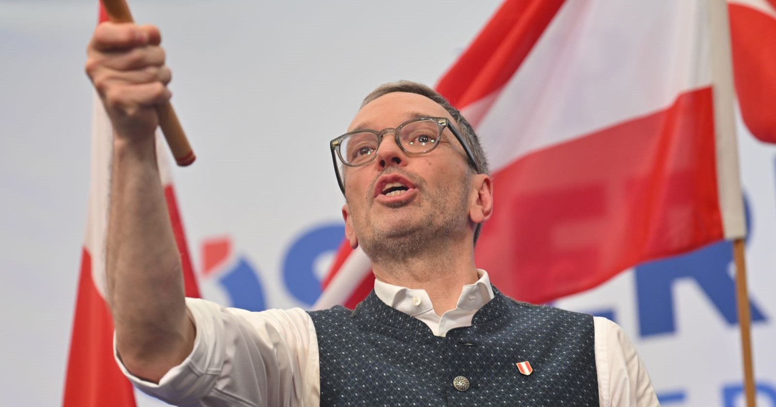 Herbert Kickl hält eine Fahne in die Höhe und spricht energisch auf der Bühne, im Hintergrund rot-weiß-rote österreichische Fahnen.