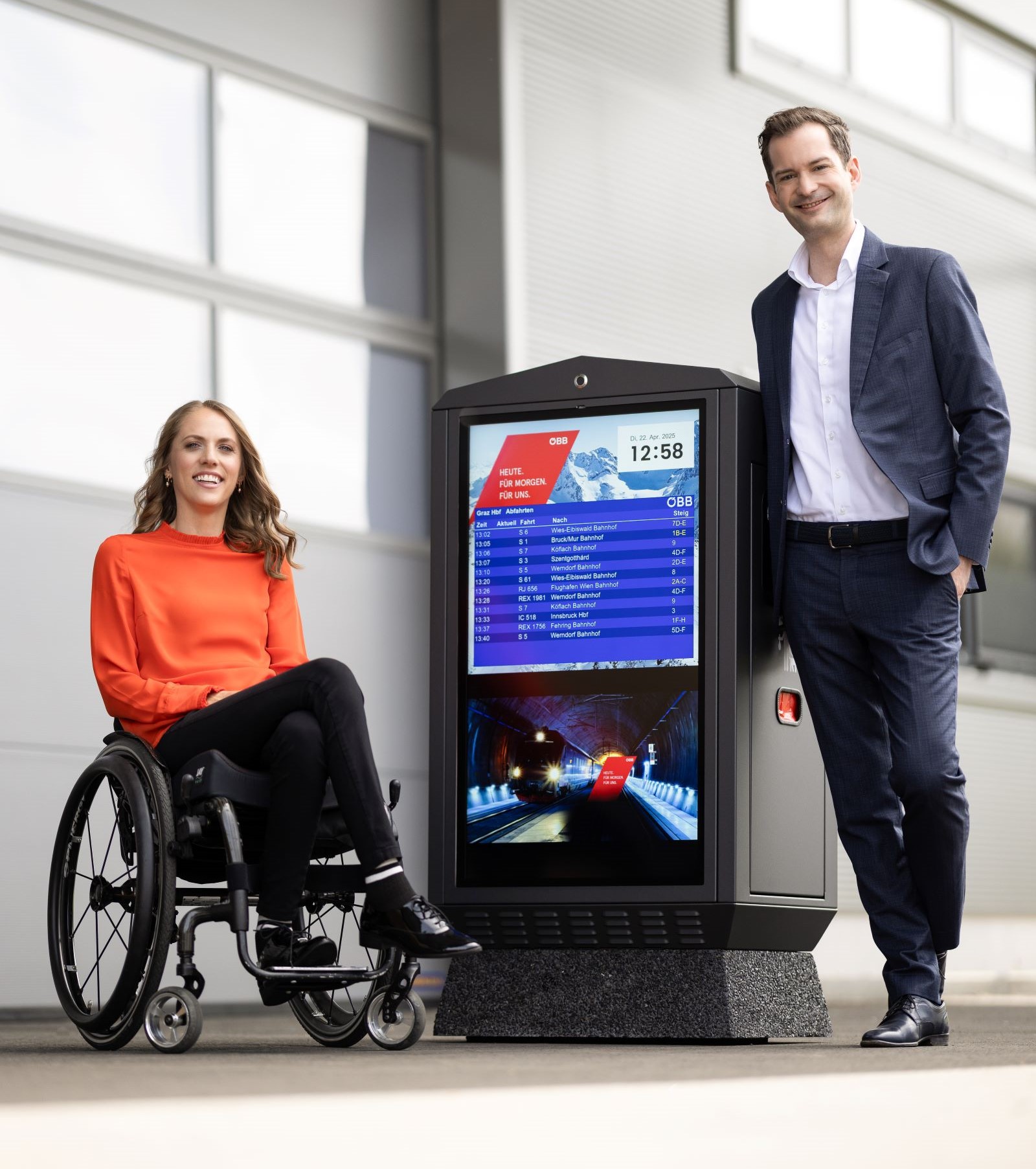 Kira Grünberg im Rollstuhl und Alexander Frank im Anzug neben einer iBox-Anzeigetafel. Die barrierefreie digitale Lösung zeigt Abfahrtszeiten und Informationen auf Augenhöhe.