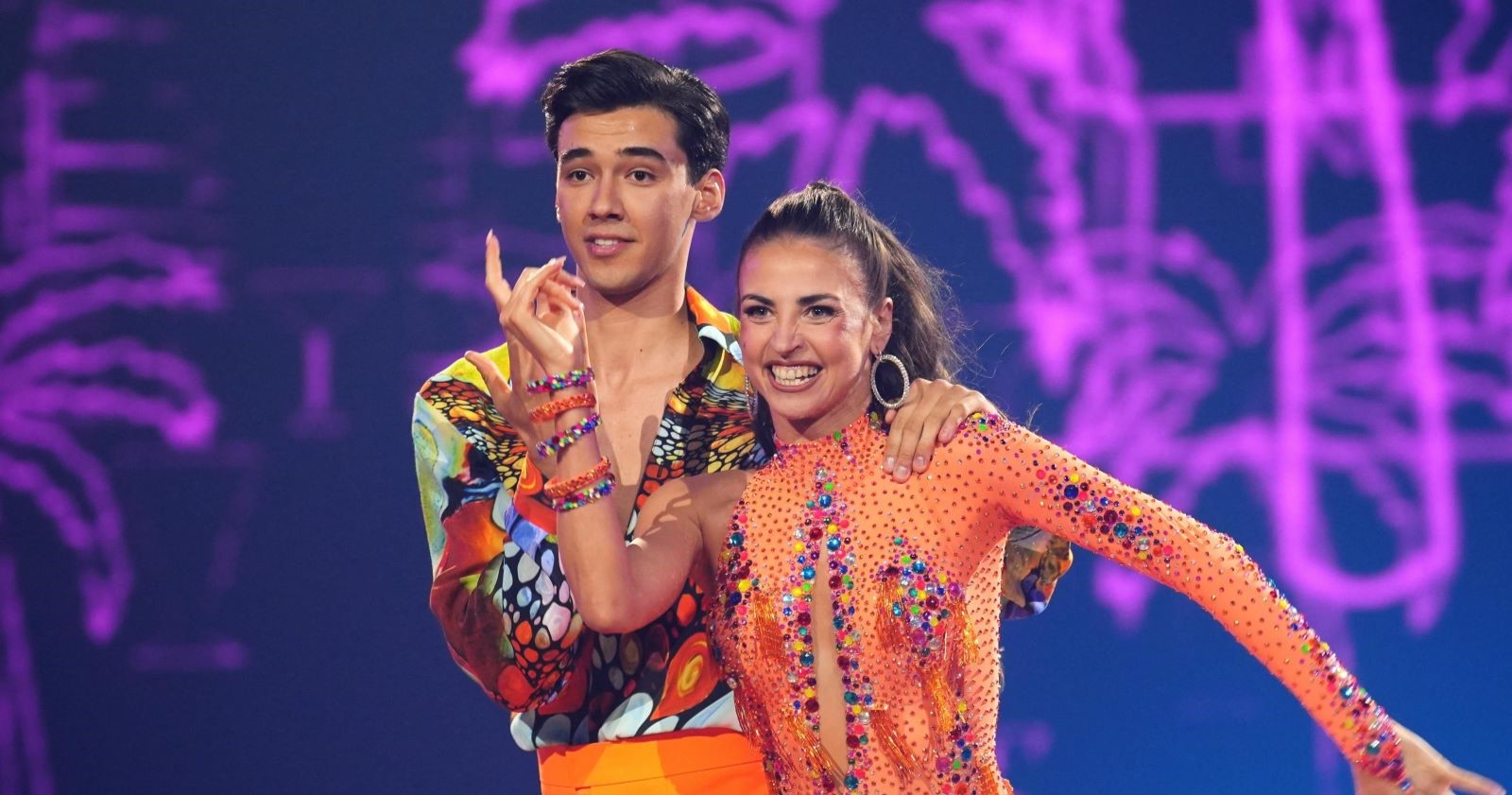 Diego Pooth und seine Tanzpartnerin Ekaterina Leonova lächeln auf der &quot;Let's Dance&quot;-Bühne im Finale 2025. Beide tragen bunte, glitzernde Kostüme, die Kulisse ist violett beleuchtet.