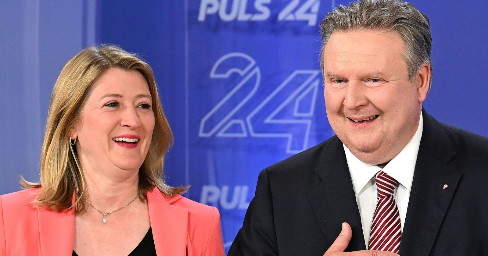Michael Ludwig (SPÖ) und Bettina Emmerling (NEOS) lachen nebeneinander bei einem TV-Auftritt vor blauem Hintergrund mit „Puls 24“-Logo – Symbolbild für die Fortsetzung der rot-pinken Koalition in Wien.