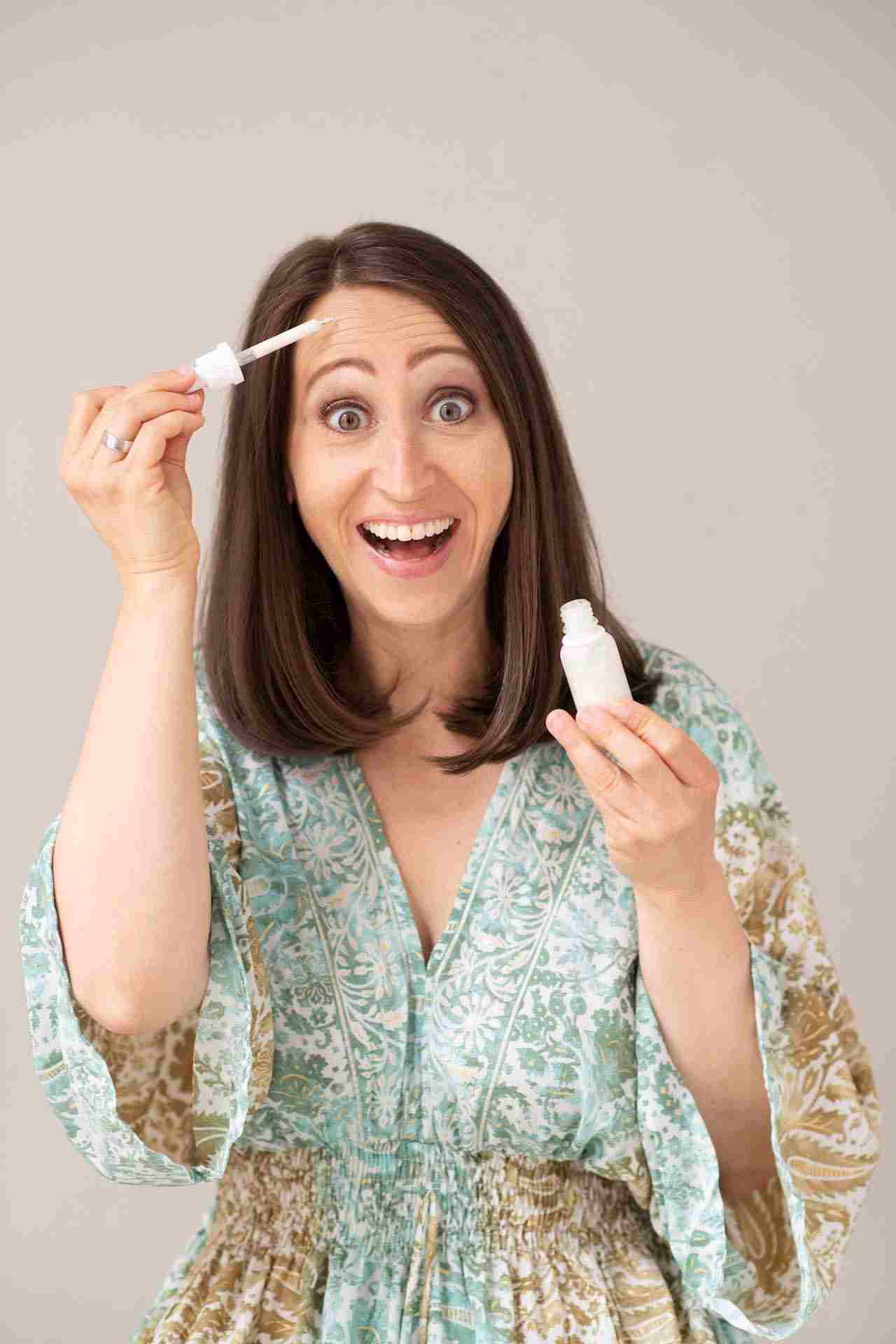 Kabarettistin Lydia Prenner-Kasper in einer türkisen, sommerhaften Bluse, in ihren Händen hält sie ein Serum, das ihre Haut verjüngen soll. Ihr Blick ist staunend, lustig.