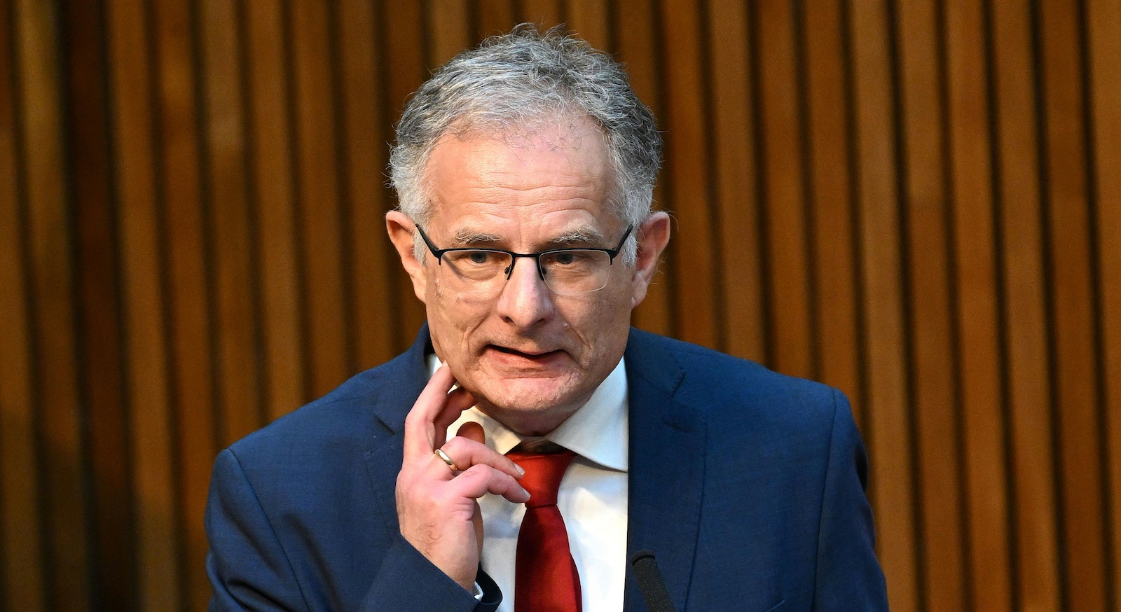 Markus Marterbauer, SPÖ-Finanzminister, spricht im Nationalrat. Er trägt einen blauen Anzug, eine rote Krawatte und eine Brille. Im Hintergrund sind Holzlamellen sichtbar.