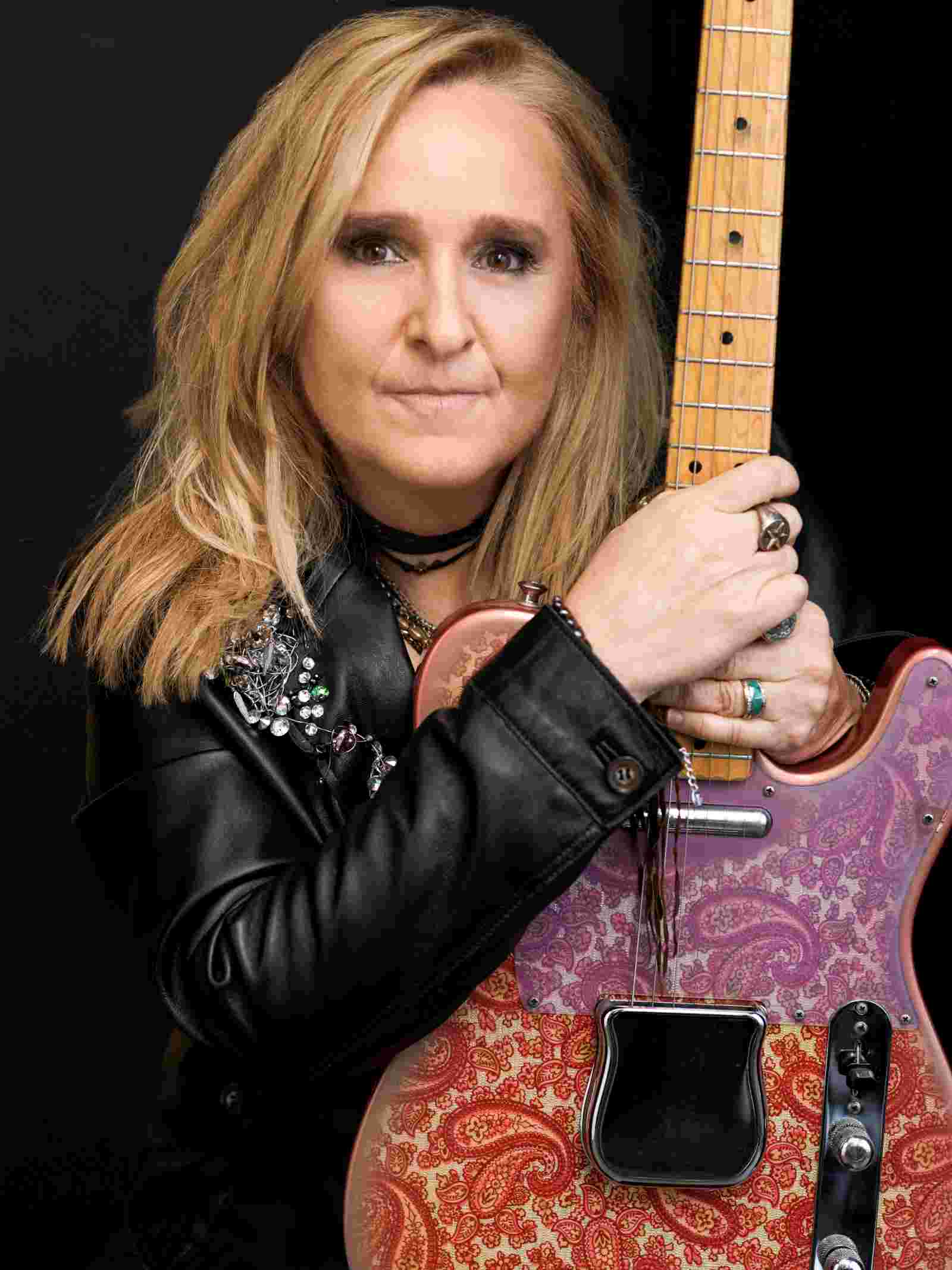 Die amerikanische Rockmusikerin Melissa Etheridge in schwarzer Lederjacke hält Gitarre auf ihrem Schoß.