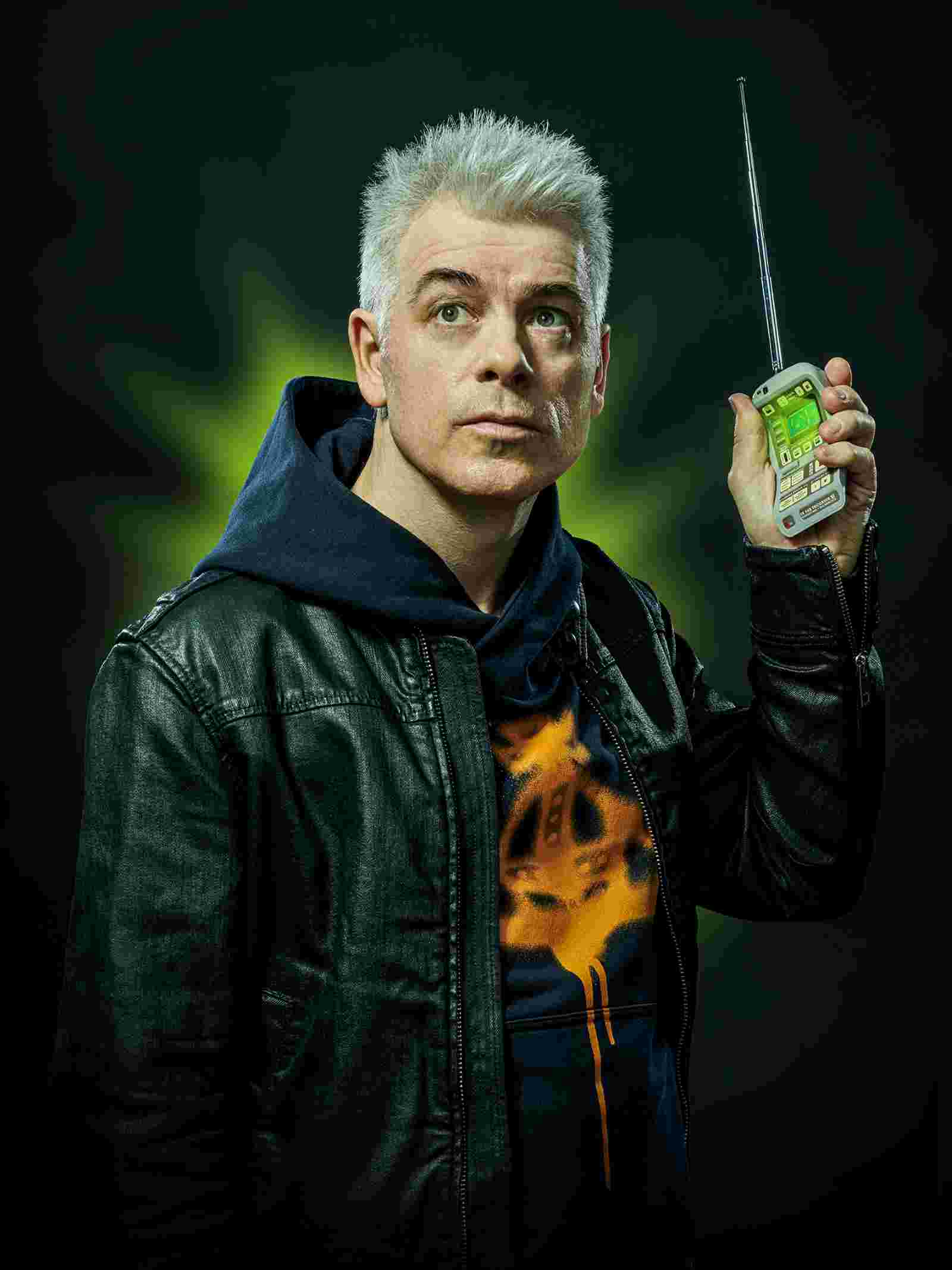 Der deutsche Kabarettist Michael Mittermeier mit grauen Haaren, trägt einen dunkelblauen Kapuzenpullover, darüber eine schwarze Lederjacke.  In seiner rechten Hand hält er ein altmodisches, silbernes Mobiltelefon mit ausziehbarer Antenne, das grün leuchtet. Er blickt leicht nach oben, sein Gesichtsausdruck ist konzentriert und nachdenklich.