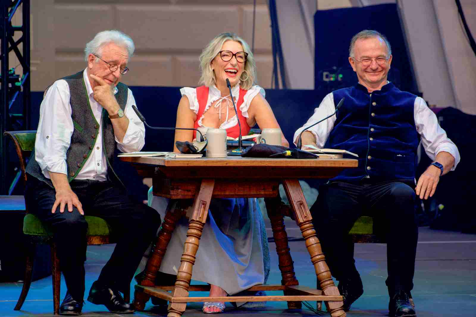 Die bayrischen Schauspielstars Gerd Anthoff, Monika Gruber und Michael Lerchenberg sitzen an einem Holztisch auf einer Bühne. Alle drei in typisch bayrischer Tracht lächeln bzw schmunzeln.