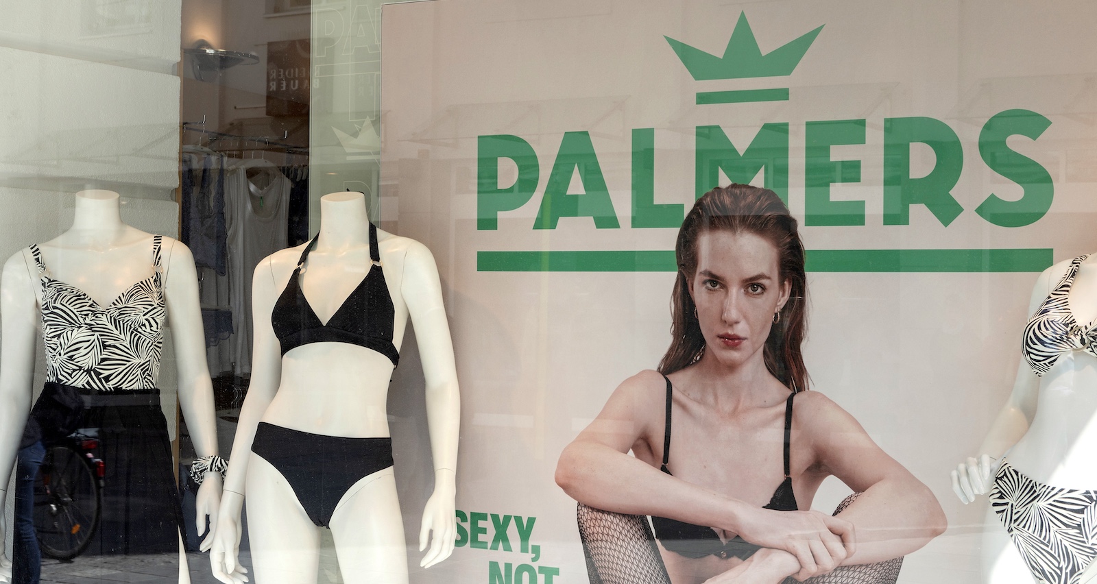 Palmers-Schaufenster mit Unterwäschemodellen und Werbeplakat einer Frau in schwarzer Lingerie, daneben das grüne Palmers-Logo.