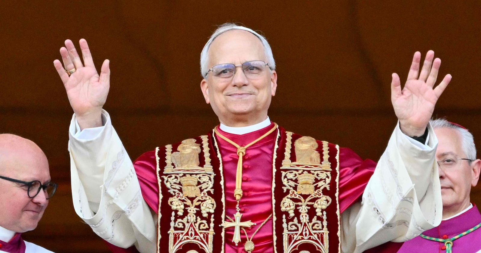 Papst Leo XIV. steht in päpstlicher liturgischer Kleidung auf dem Balkon des Petersdoms und hebt beide Hände zum Gruß. Links und rechts neben ihm stehen Kirchenvertreter.