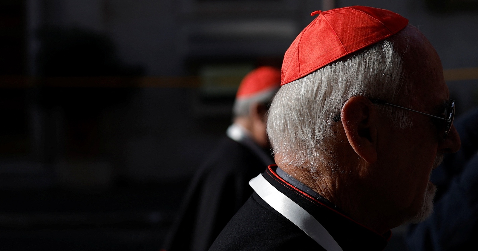 Ein älterer Kardinal mit rotem Birett und schwarzer Soutane mit roten Paspeln steht im Profil. Im Hintergrund ist ein weiterer Kardinal unscharf zu erkennen. Die Szene spielt sich im Zwielicht auf einer Straße ab.