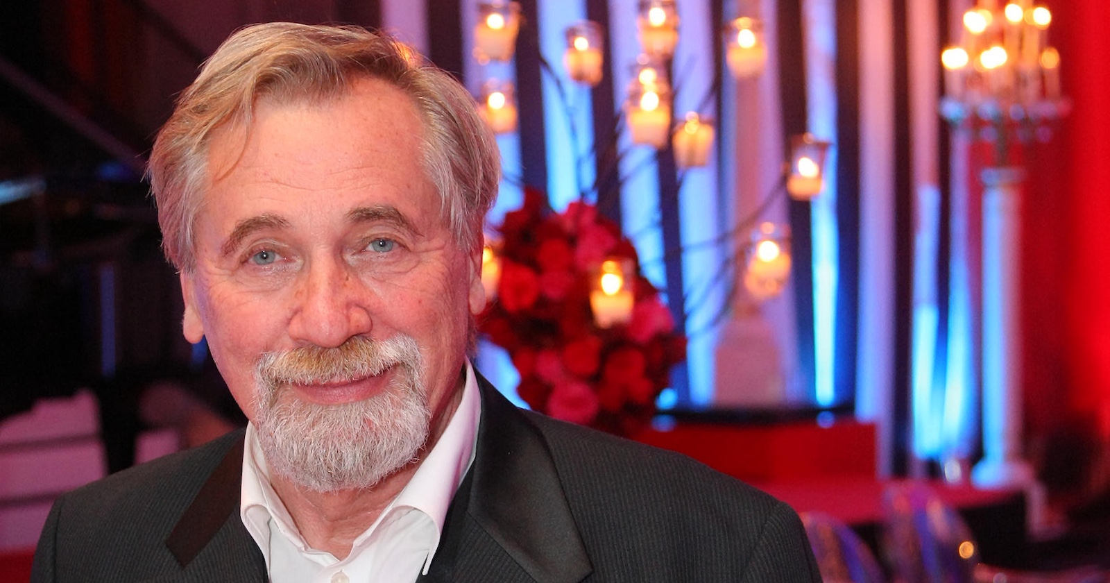 Porträt von Peter Rapp mit weißem Haar und Bart, freundlich lächelnd in festlichem Ambiente mit Kerzenlichtern und rotem Blumenschmuck im Hintergrund.